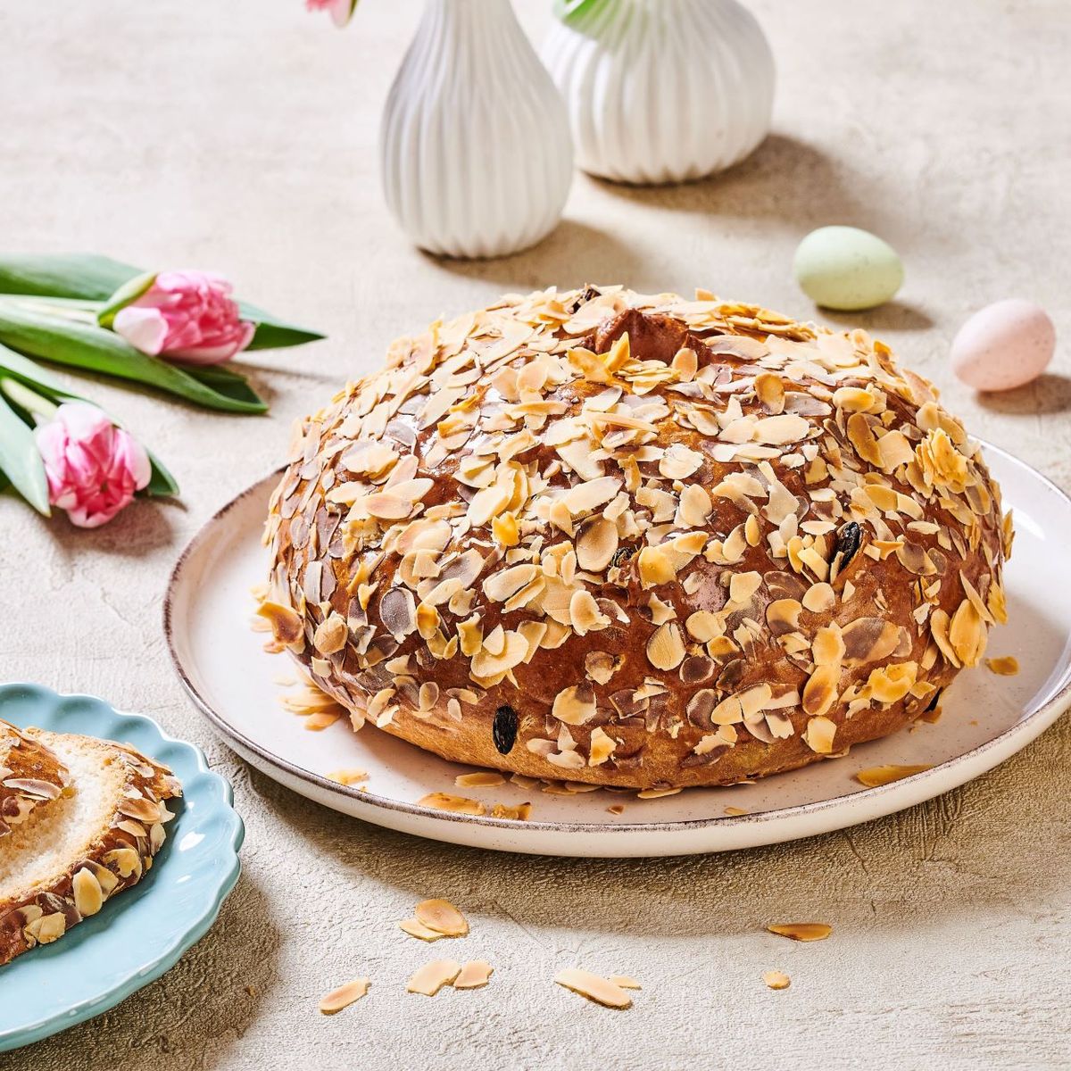 Kulatý mandlový dort na talíři, obklopený tulipány a malými vajíčky, a jeho nakrájená verze na modrém talíři.