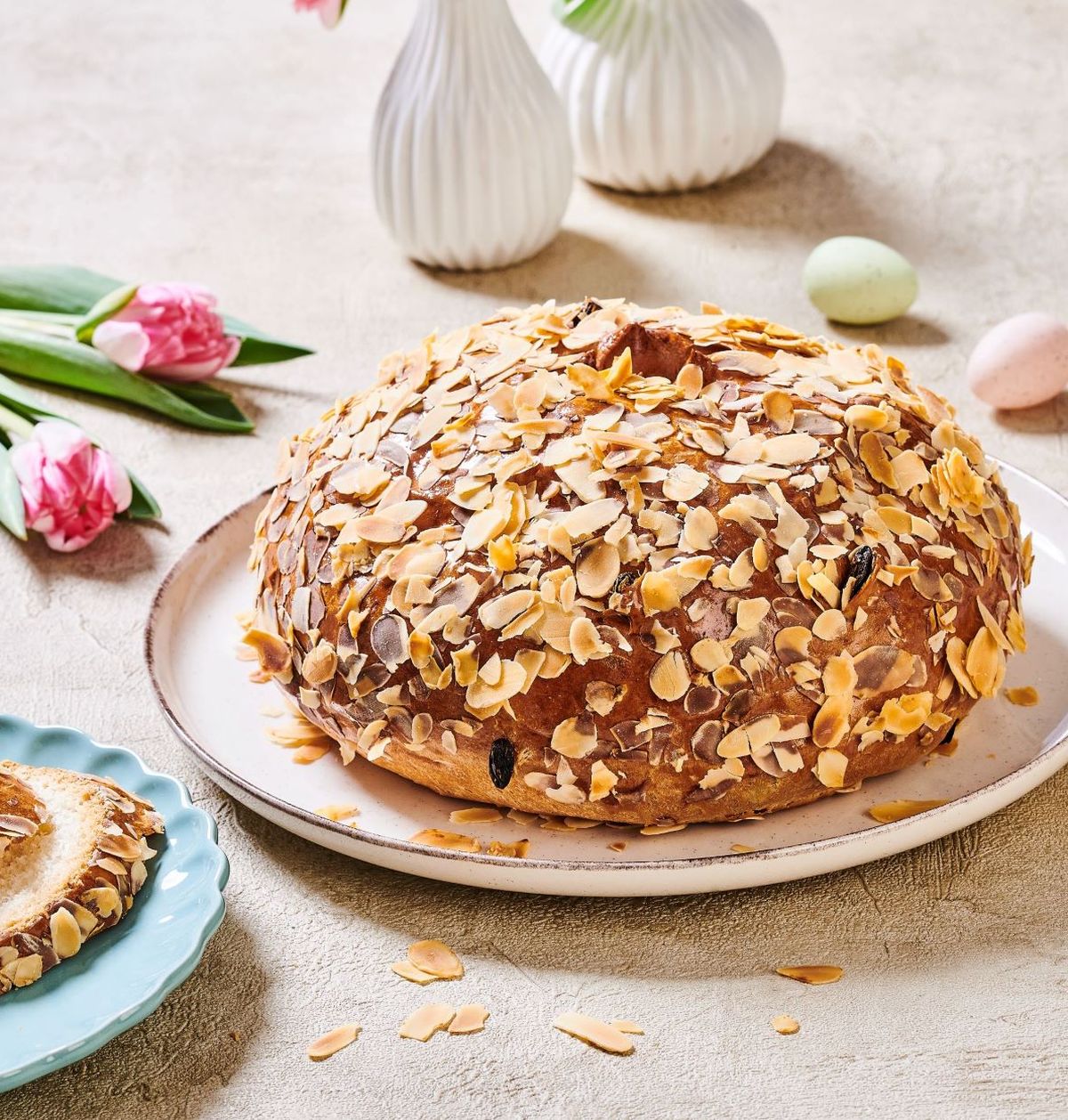 Kulatý mandlový dort na talíři, obklopený tulipány a malými vajíčky, a jeho nakrájená verze na modrém talíři.