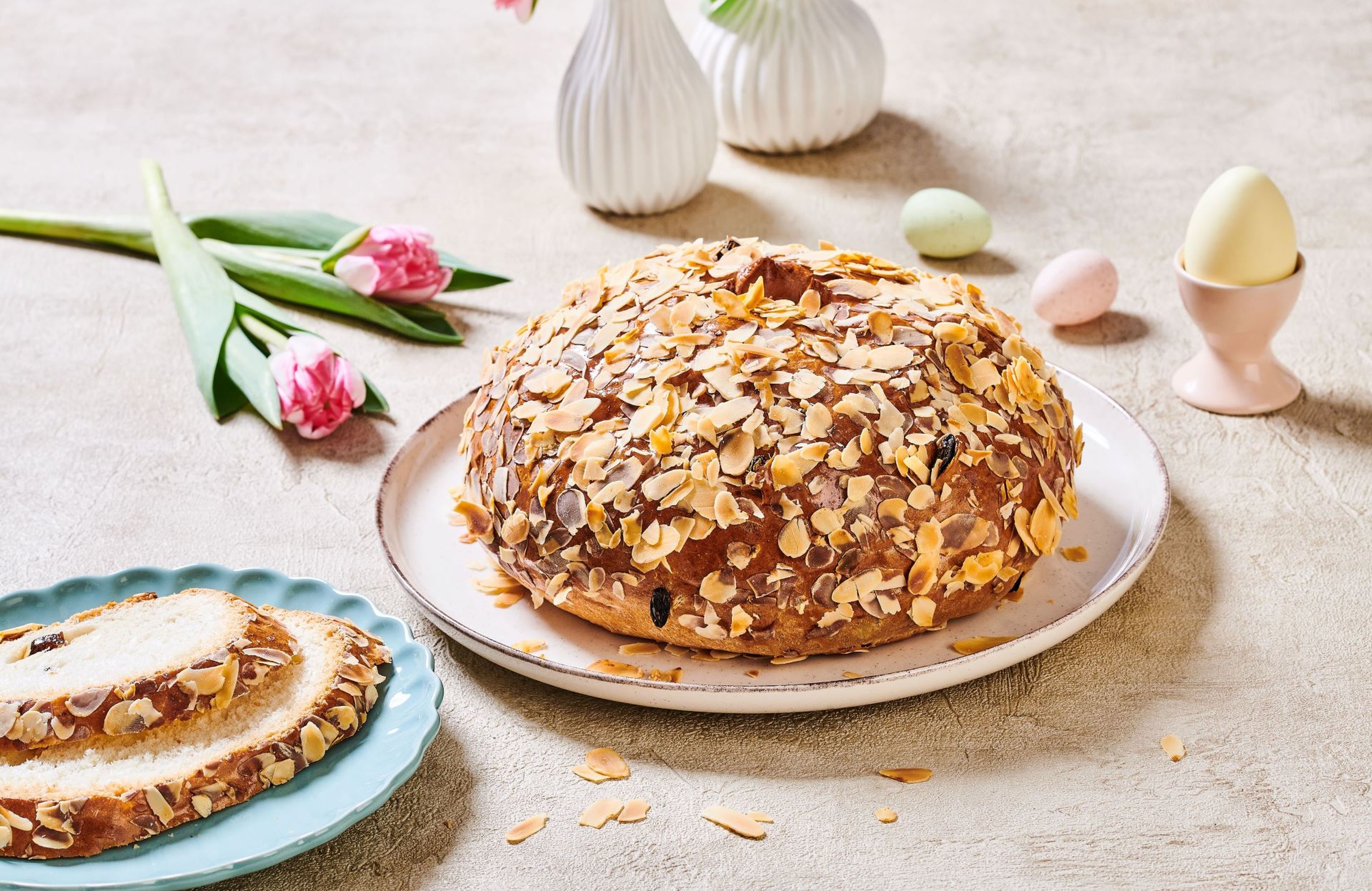 Kulatý mandlový dort na talíři, obklopený tulipány a malými vajíčky, a jeho nakrájená verze na modrém talíři.