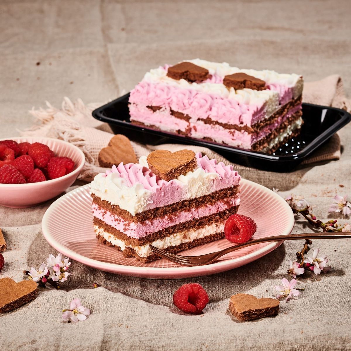 Ružovo-biela vrstvená torta so srdcovými sušienkami, obklopená malinami a kvetmi na textúrovanom látkovom pozadí.