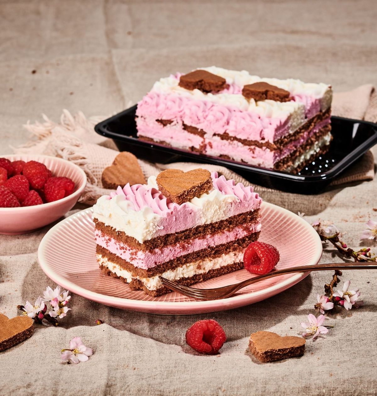 Ružovo-biela vrstvená torta so srdcovými sušienkami, obklopená malinami a kvetmi na textúrovanom látkovom pozadí.
