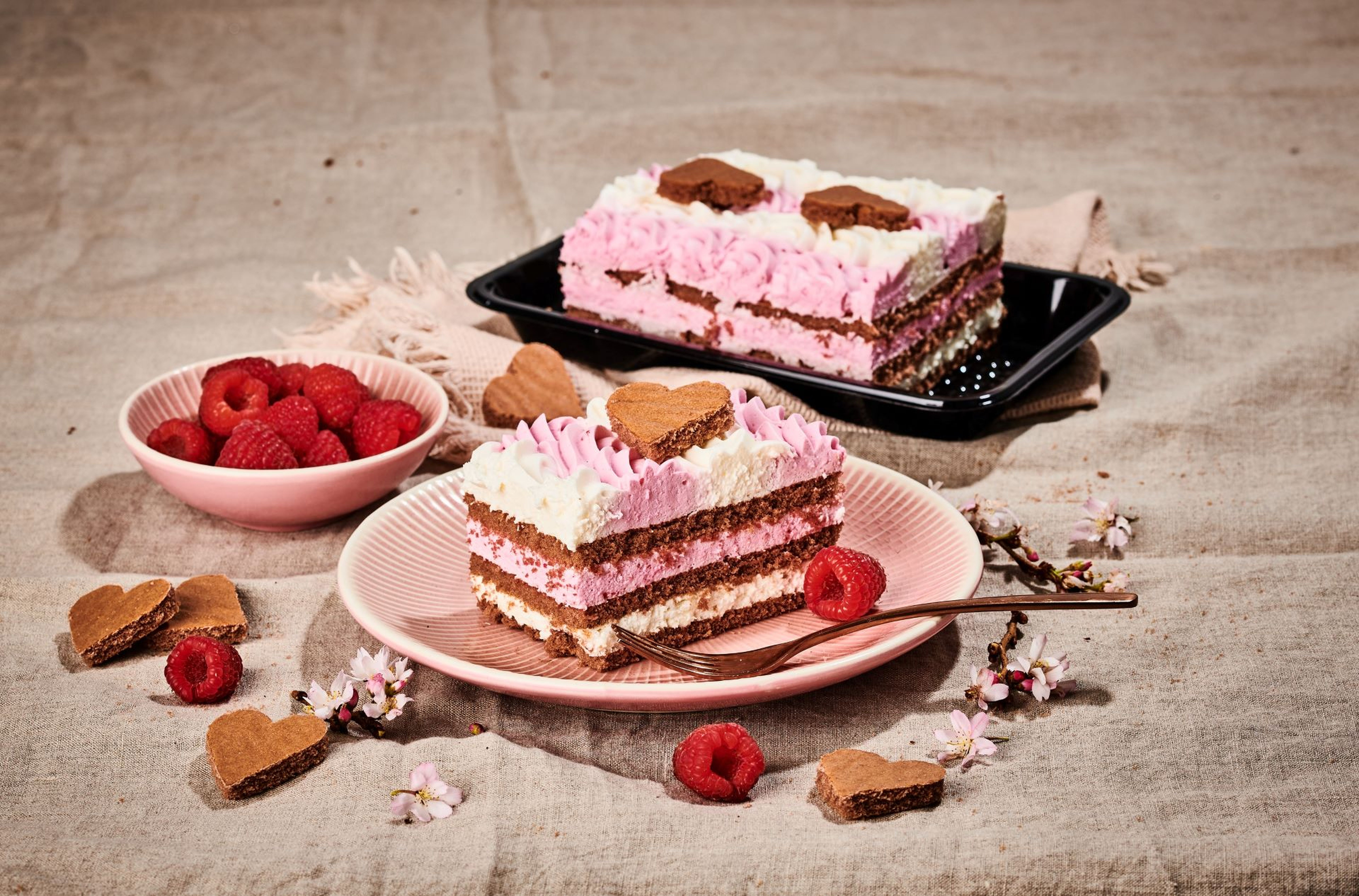 Ružovo-biela vrstvená torta so srdcovými sušienkami, obklopená malinami a kvetmi na textúrovanom látkovom pozadí.