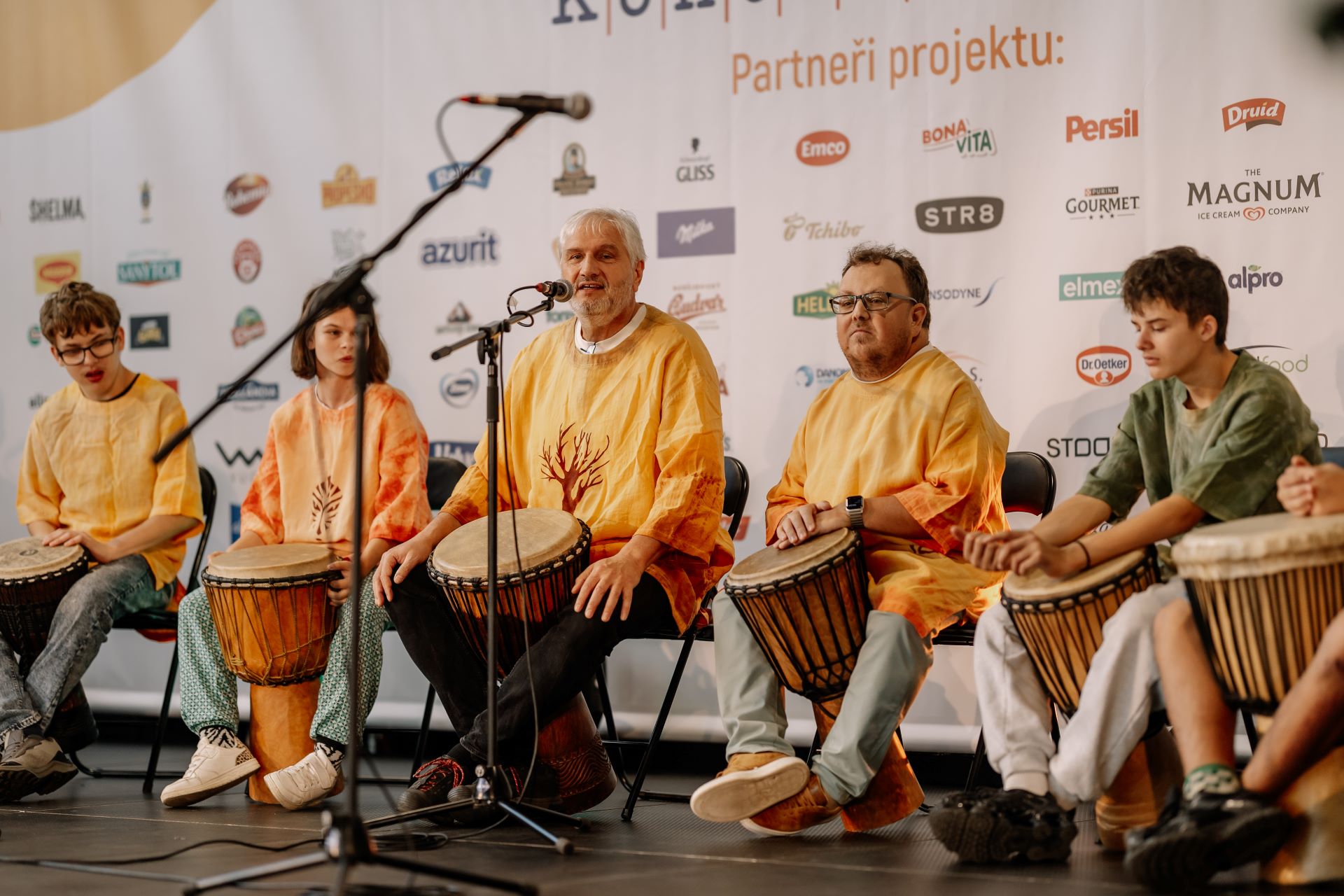 Skupina lidí v oranžových tričkách, kteří na jevišti hrají na ruční bubny, za nimi je na pozadí vidět několik log.