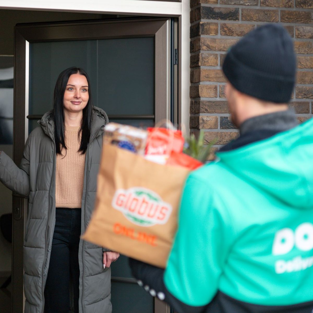 Žena se usmívá, když přebírá zásilku s potravinami před svým domem. Doručovatel má na sobě zelenou bundu s nápisem „Dodo“ a drží papírovou tašku.
