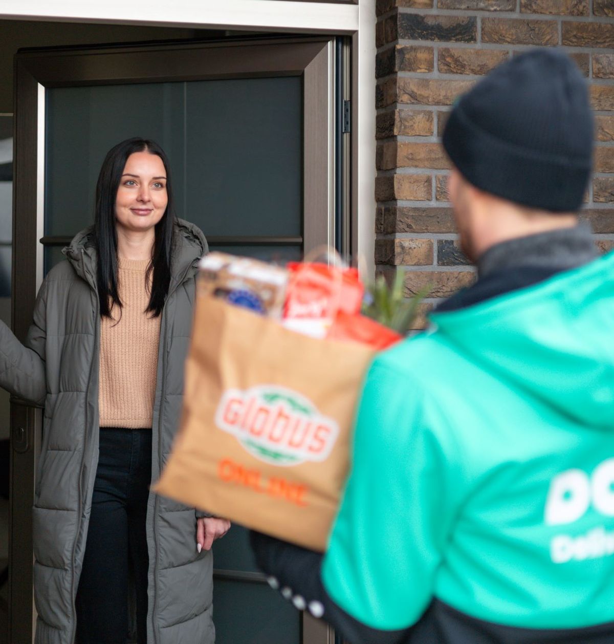 Žena se usmívá, když přebírá zásilku s potravinami před svým domem. Doručovatel má na sobě zelenou bundu s nápisem „Dodo“ a drží papírovou tašku.