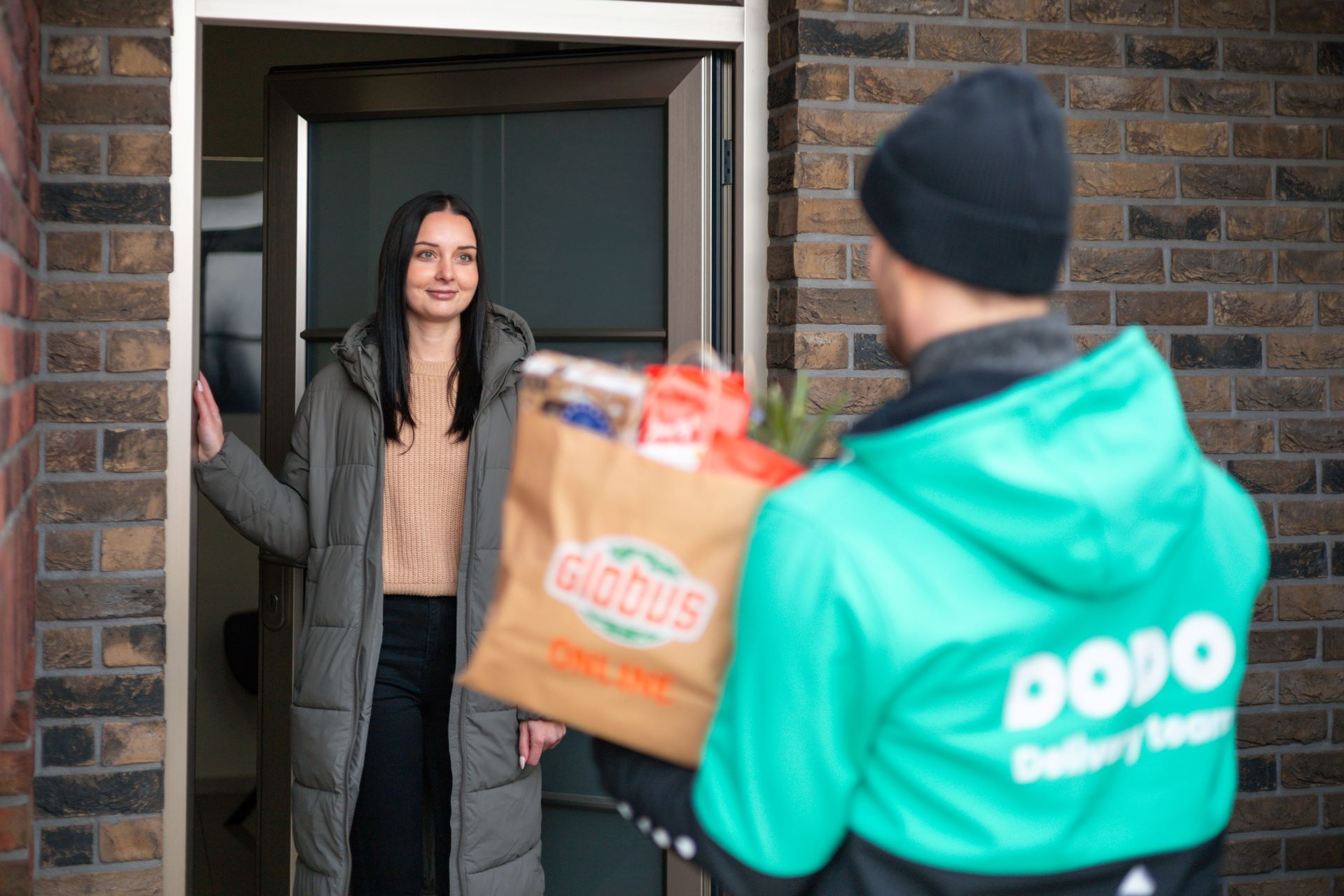 Žena se usmívá, když přebírá zásilku s potravinami před svým domem. Doručovatel má na sobě zelenou bundu s nápisem „Dodo“ a drží papírovou tašku.