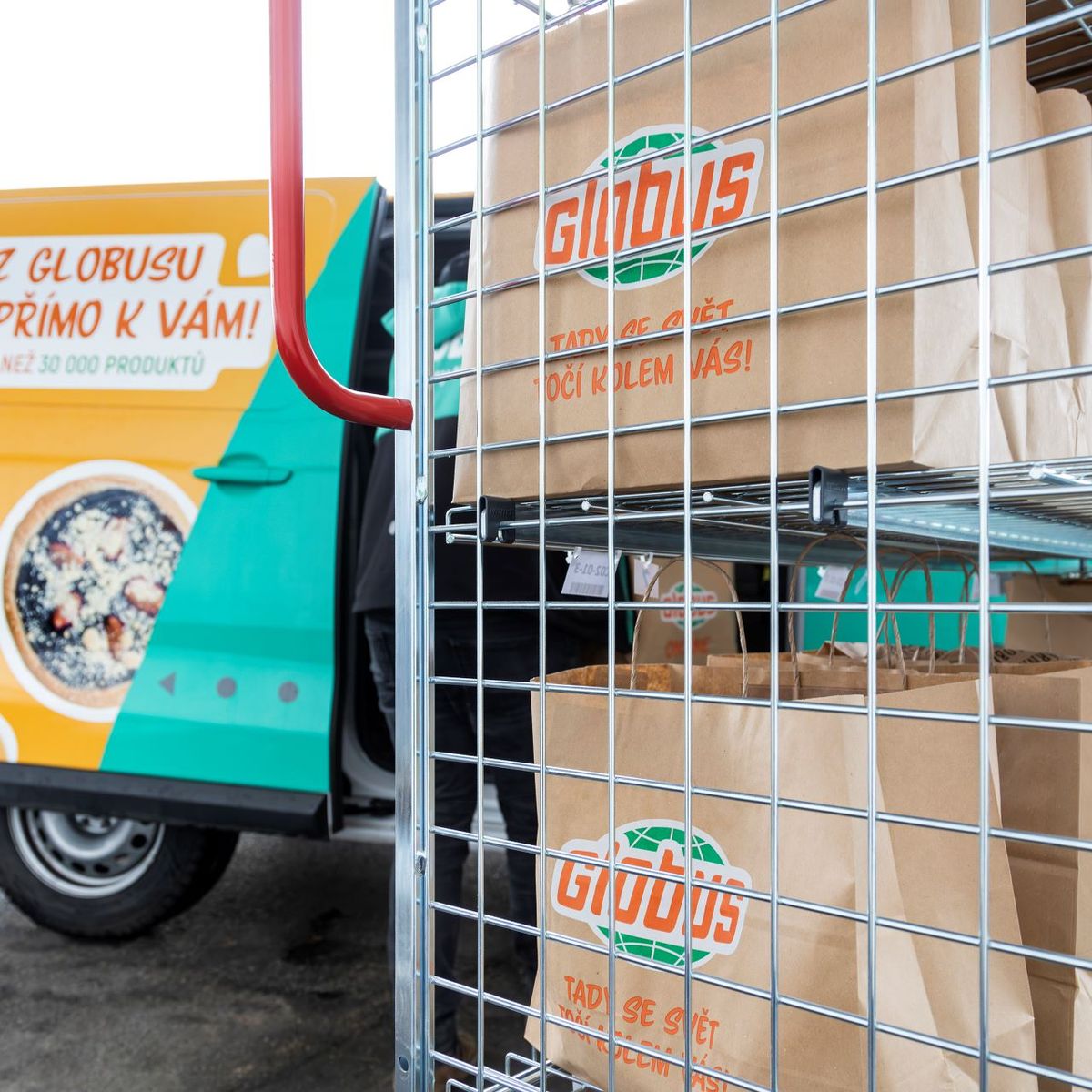 Dodávka s logem „Globus“ stojí s otevřenými dveřmi vedle kovových regálů zaplněných hnědými papírovými taškami, které jsou zřejmě určeny k rozvozu potravin.