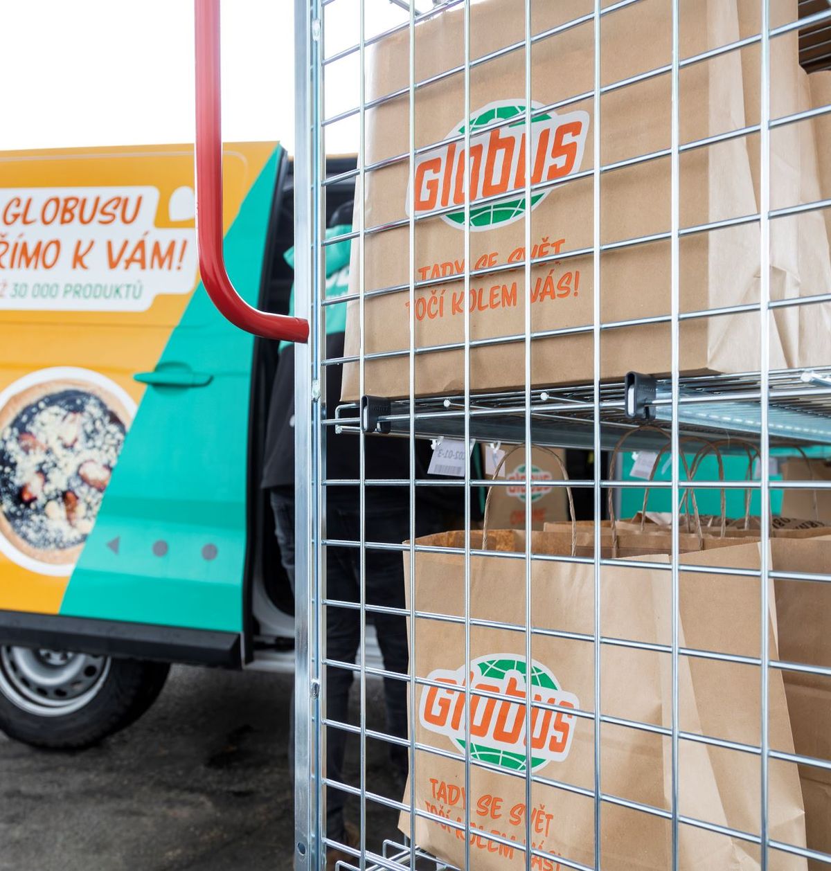 Dodávka s logem „Globus“ stojí s otevřenými dveřmi vedle kovových regálů zaplněných hnědými papírovými taškami, které jsou zřejmě určeny k rozvozu potravin.