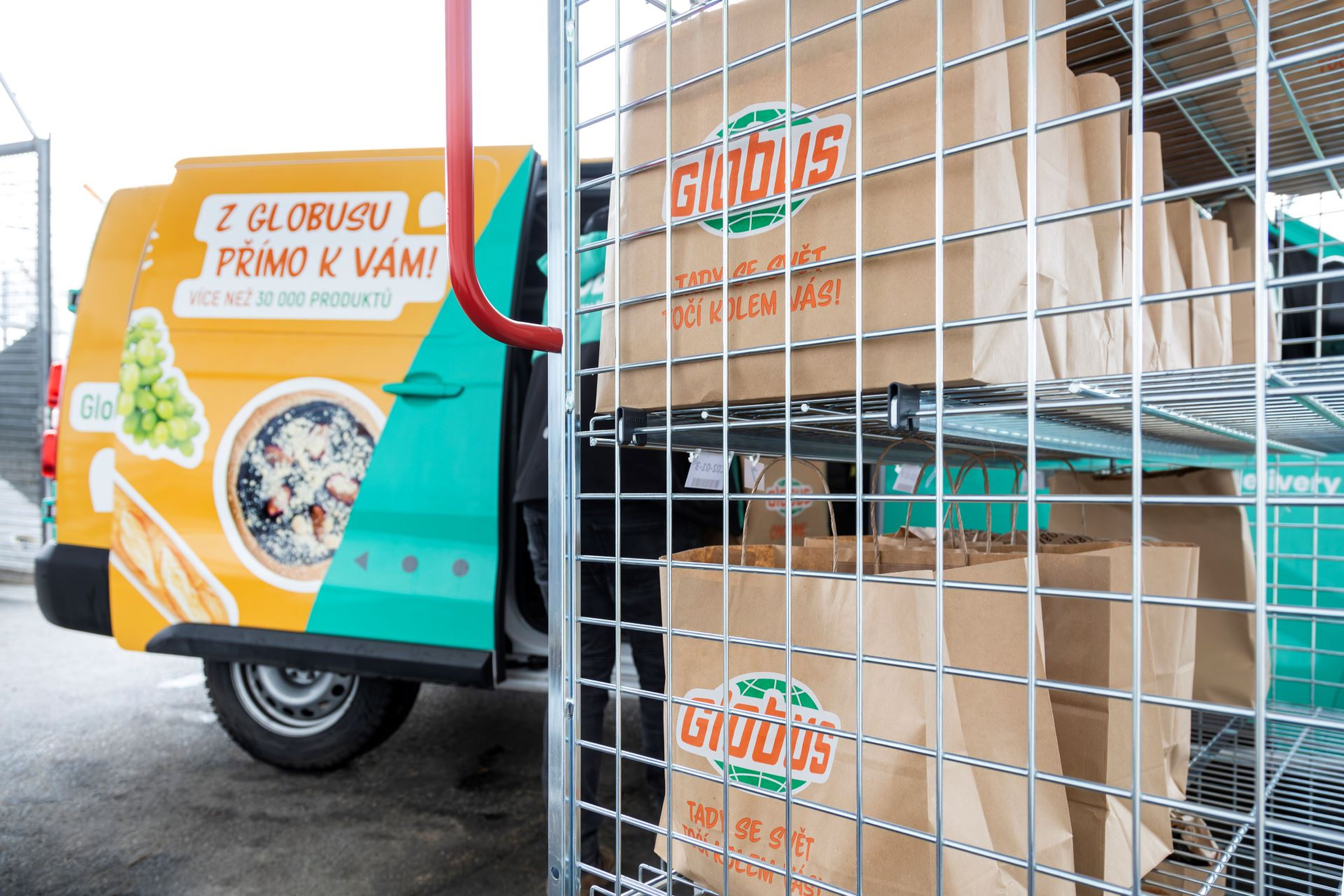 Dodávka s logem „Globus“ stojí s otevřenými dveřmi vedle kovových regálů zaplněných hnědými papírovými taškami, které jsou zřejmě určeny k rozvozu potravin.