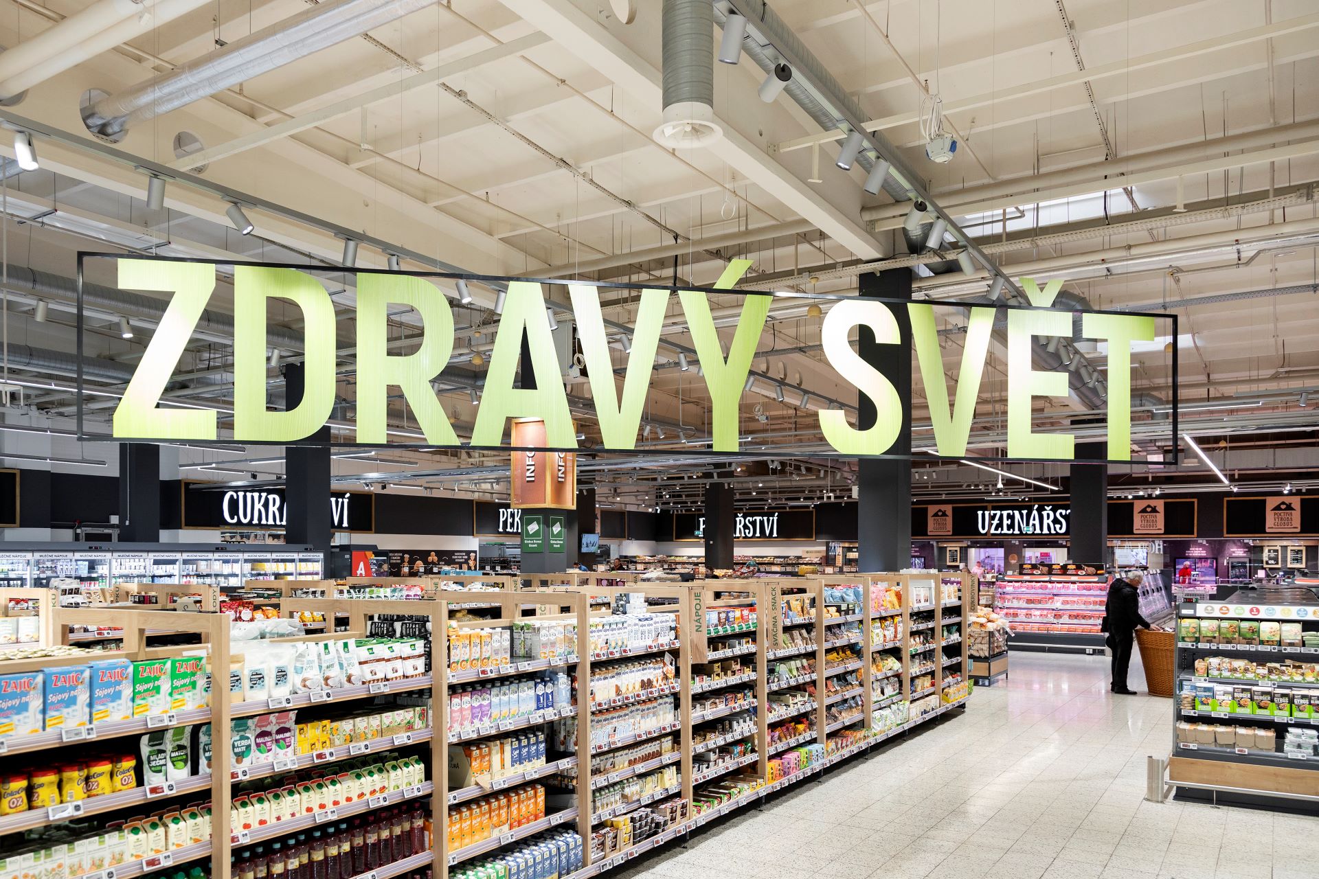 Široká ulička supermarketu s regály s potravinami pod velkým nápisem „ZDRAVÝ SVĚT“ v prostorném, dobře osvětleném interiéru.