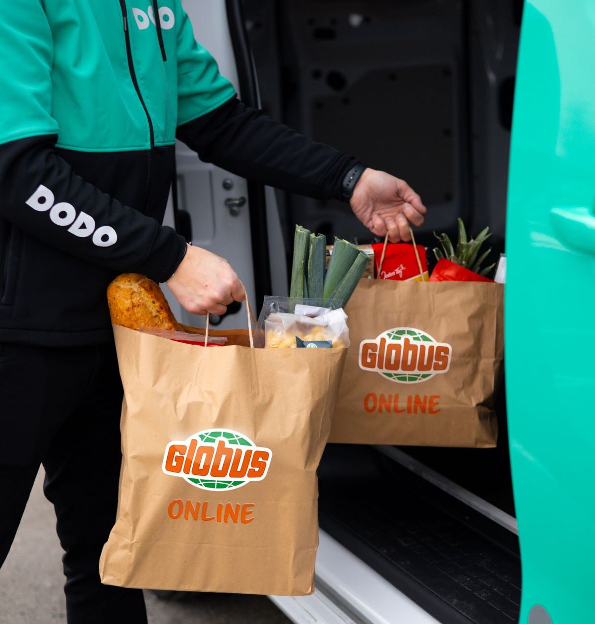 Osoba vykládající z dodávky tašky s potravinami s nápisem „Globus Online“, které obsahují chléb, zeleninu a další potraviny.