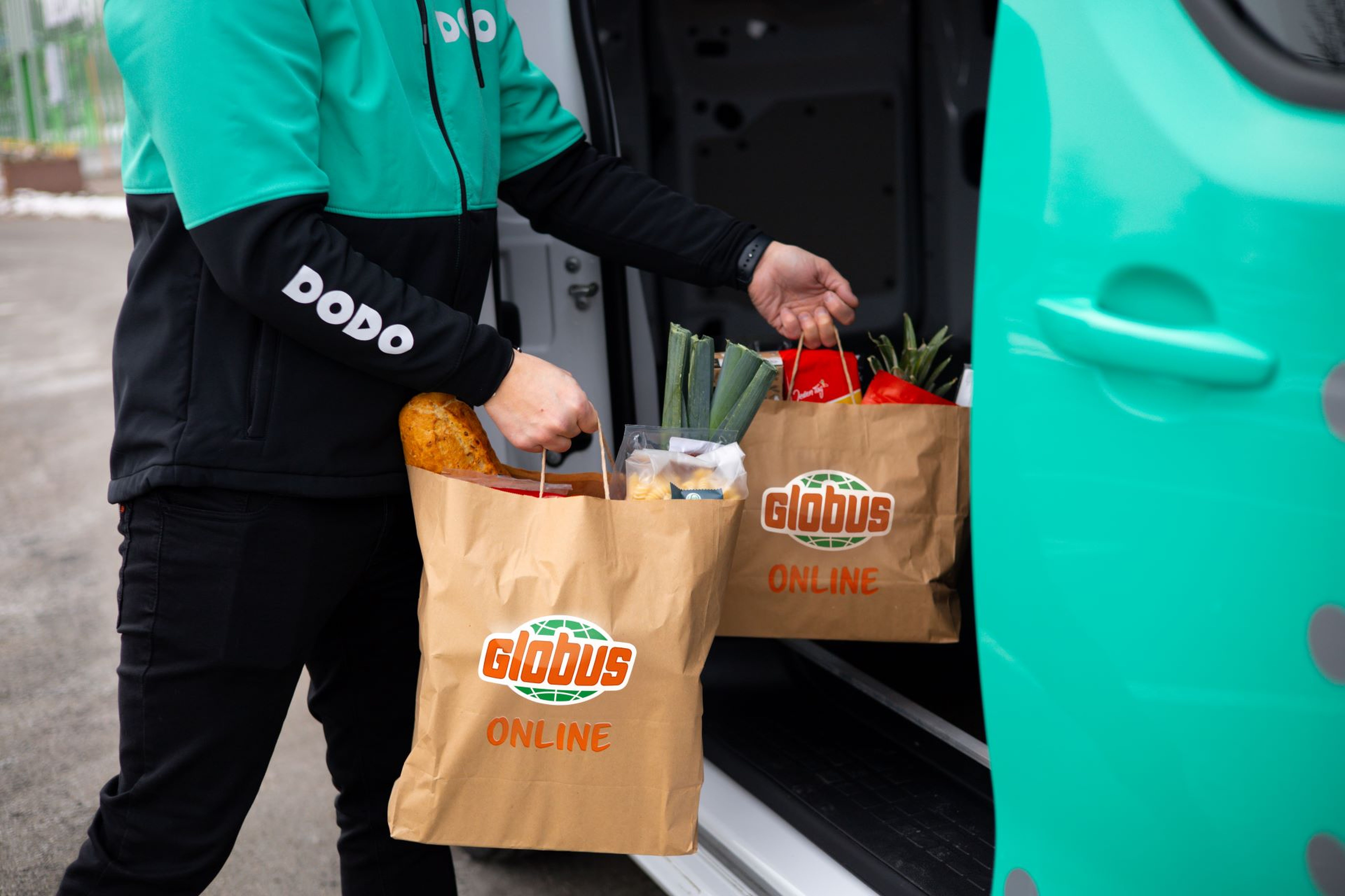 Osoba vykládající z dodávky tašky s potravinami s nápisem „Globus Online“, které obsahují chléb, zeleninu a další potraviny.