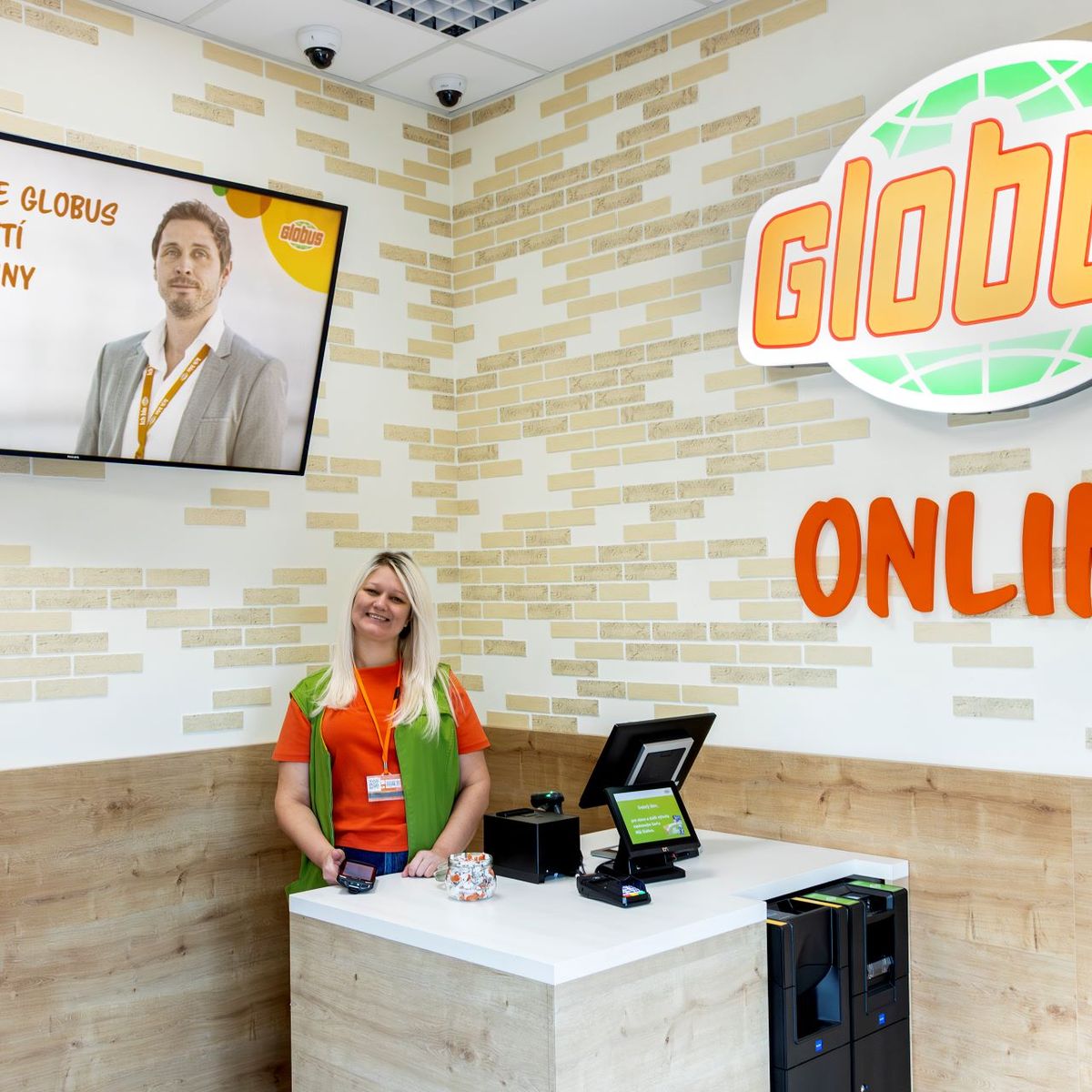 Usměvavý zaměstnanec u přepážky Globus Online s digitálním displejem a logem na zdi.