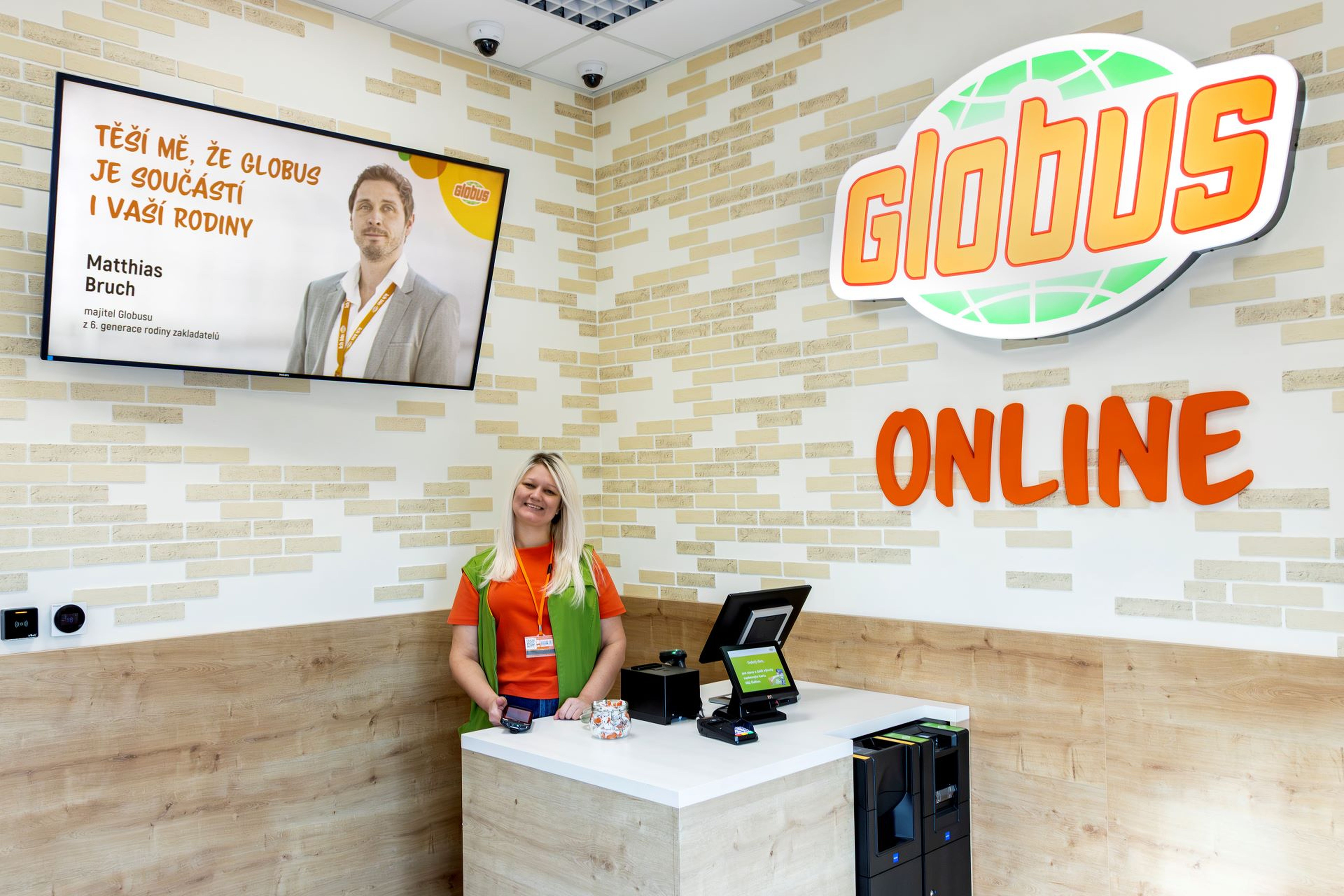 Usměvavý zaměstnanec u přepážky Globus Online s digitálním displejem a logem na zdi.