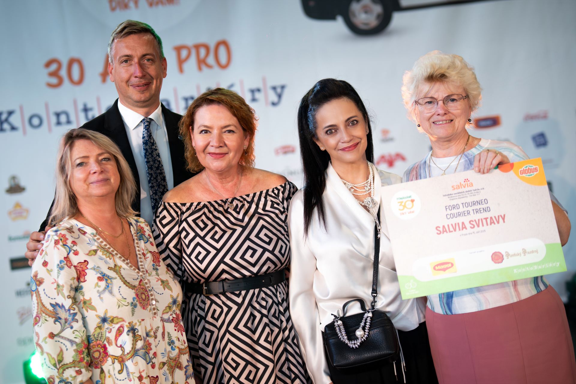 Pět lidí pózuje společně, z nichž jeden drží velký certifikát. V pozadí je vidět branding akce a loga.