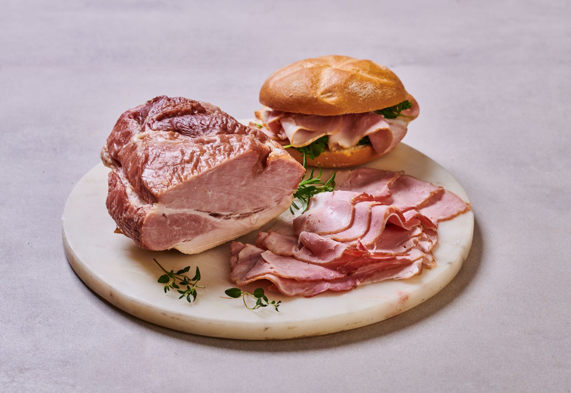 Nakrájená šunka na mramorové desce s bylinkami, vedle sendviče se šunkou a zeleninou v housce.