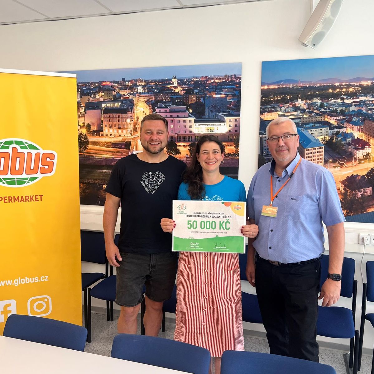 Tři lidé stojí v interiéru a drží velký šek na 50 000 Kč. V pozadí je banner „Globus Hypermarket“ a fotografie městské krajiny.