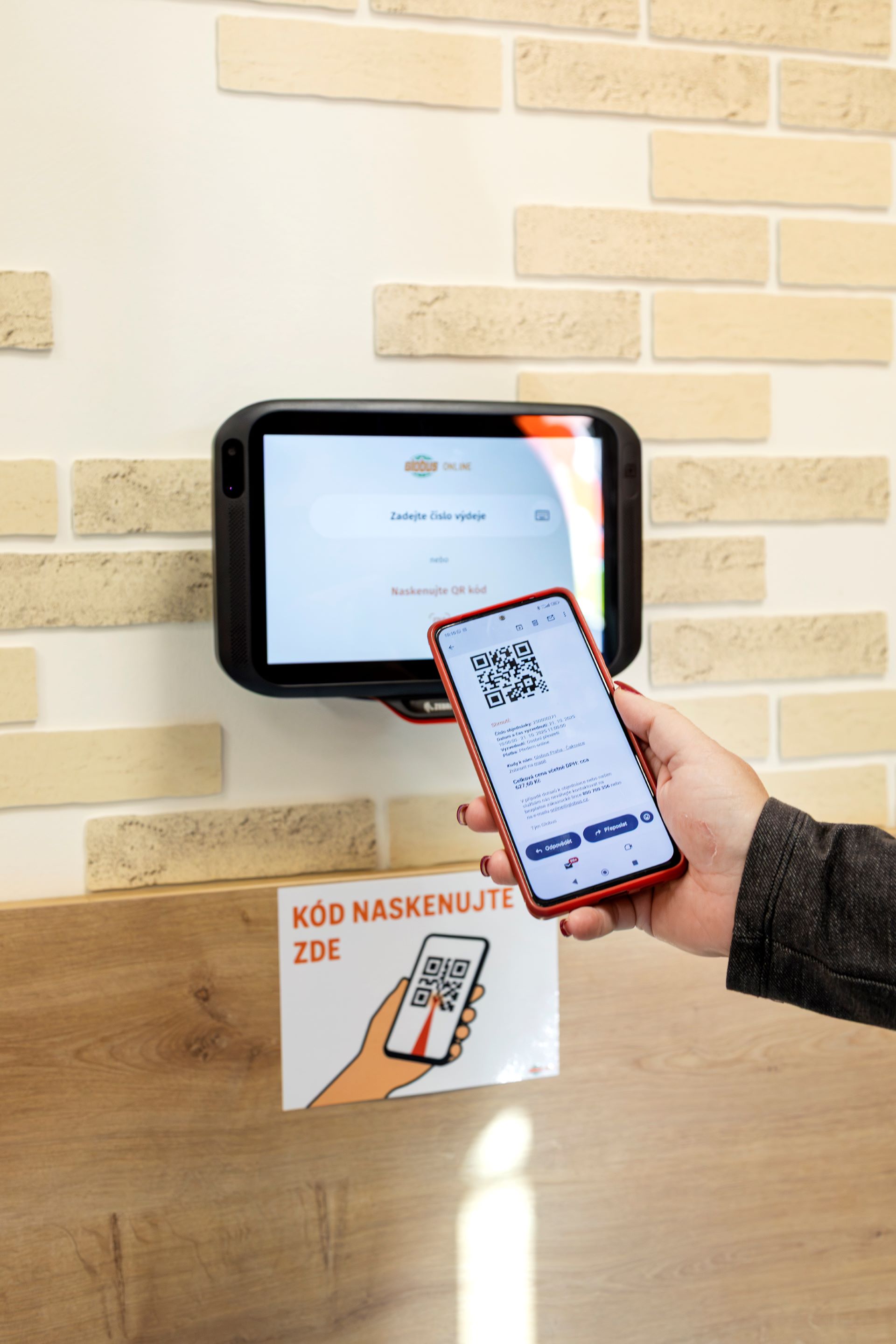 Osoba skenuje QR kód na smartphonu před tabletem připevněným na zdi. Pod ním je nápis „KÓD NASKENUJTE ZDE“ s ikonou QR kódu.