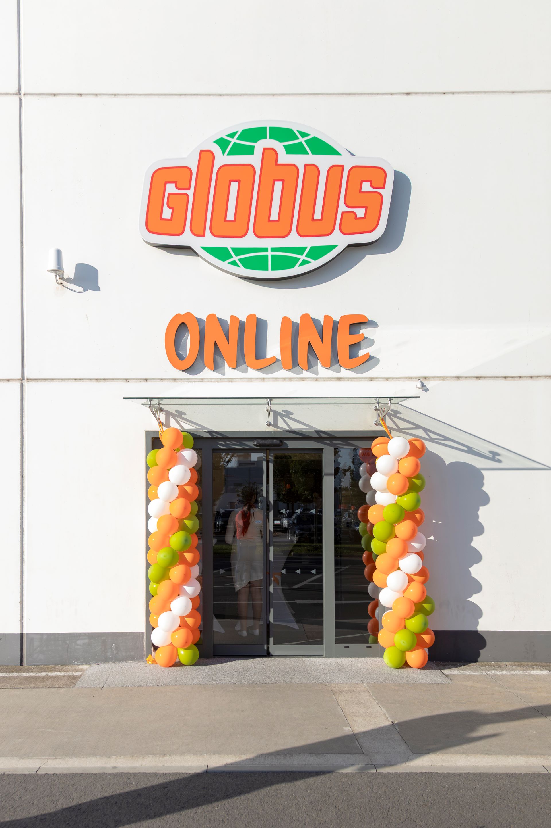 Výloha s nápisem „Globus Online“ nad vchodem, zdobená oranžovými, bílými a zelenými balónky po obou stranách skleněných dveří.