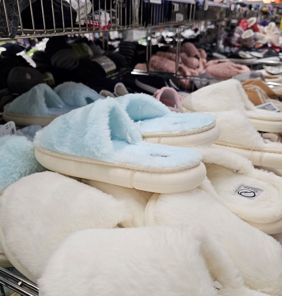 Regál v obchodě plný různých měkkých pantoflí v pastelových barvách, včetně modré a bílé, uspořádaných v úhledných řadách.