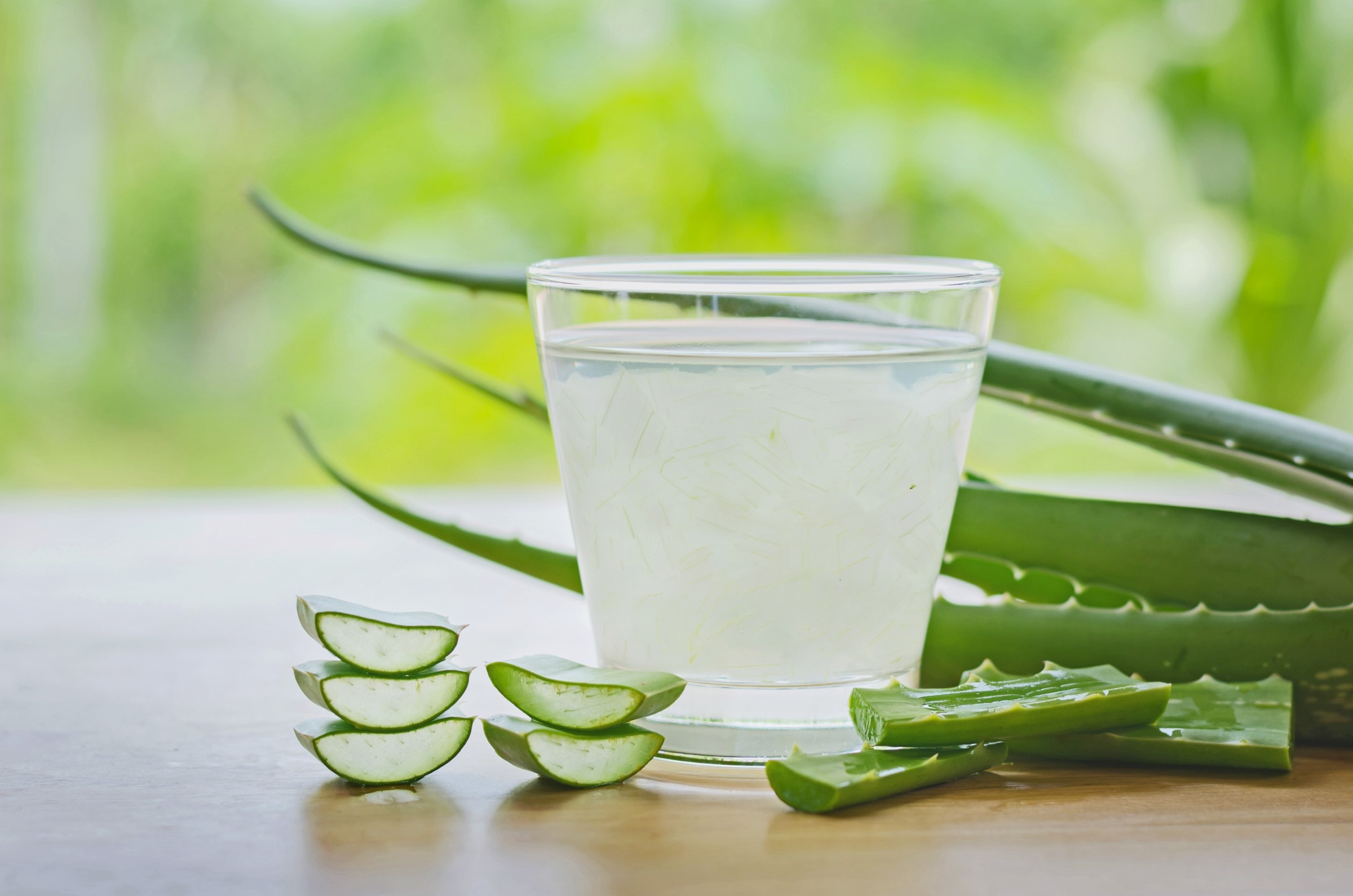 Sklenice průzračného aloe vera džusu vedle naskládaných nakrájených kousků aloe a listů na dřevěné ploše s jemně zeleným pozadím.