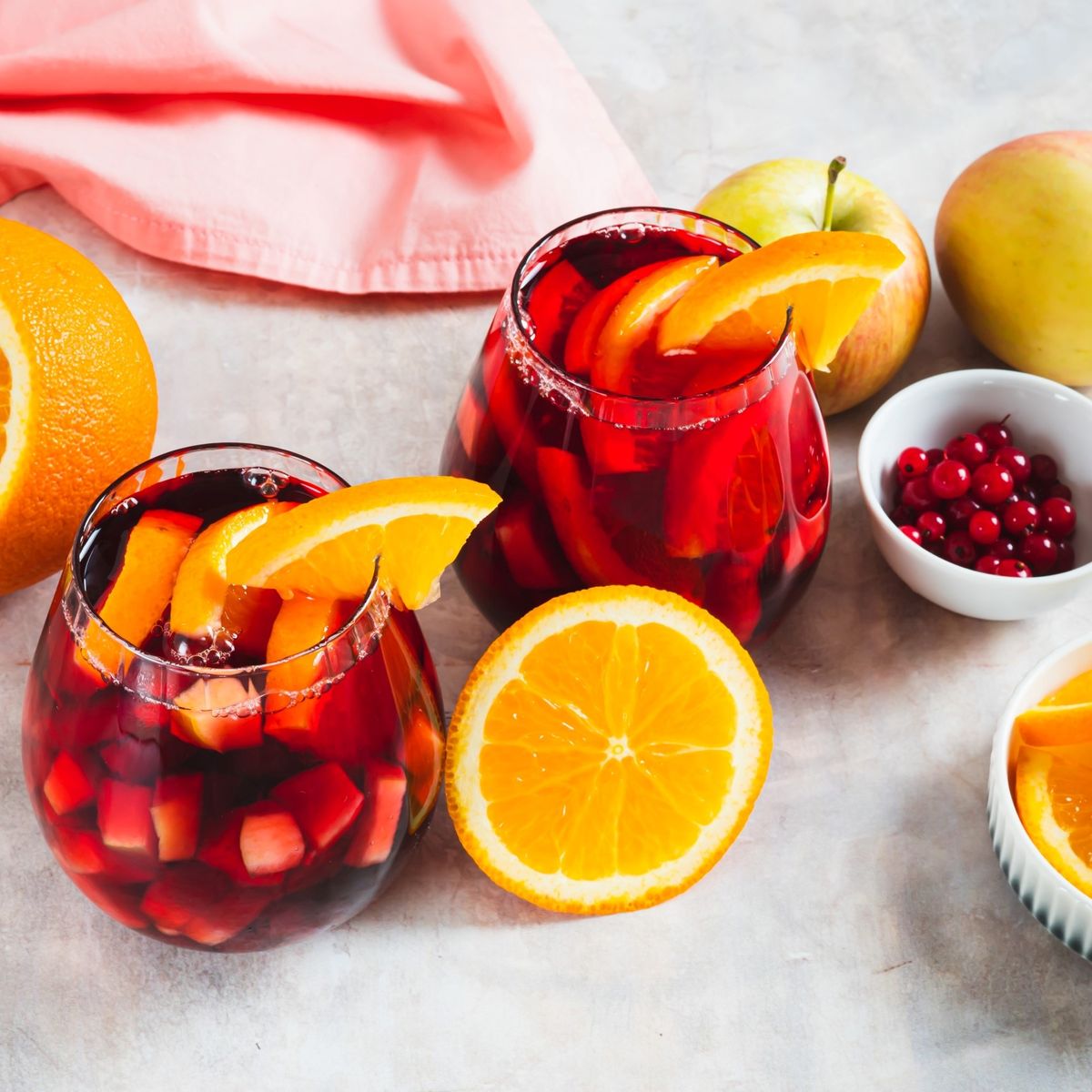 Dvě sklenice červené sangrie s plátky pomeranče, obklopené čerstvými pomeranči, jablky, růžovým ubrusem, miskou brusinek a kousky pomeranče.