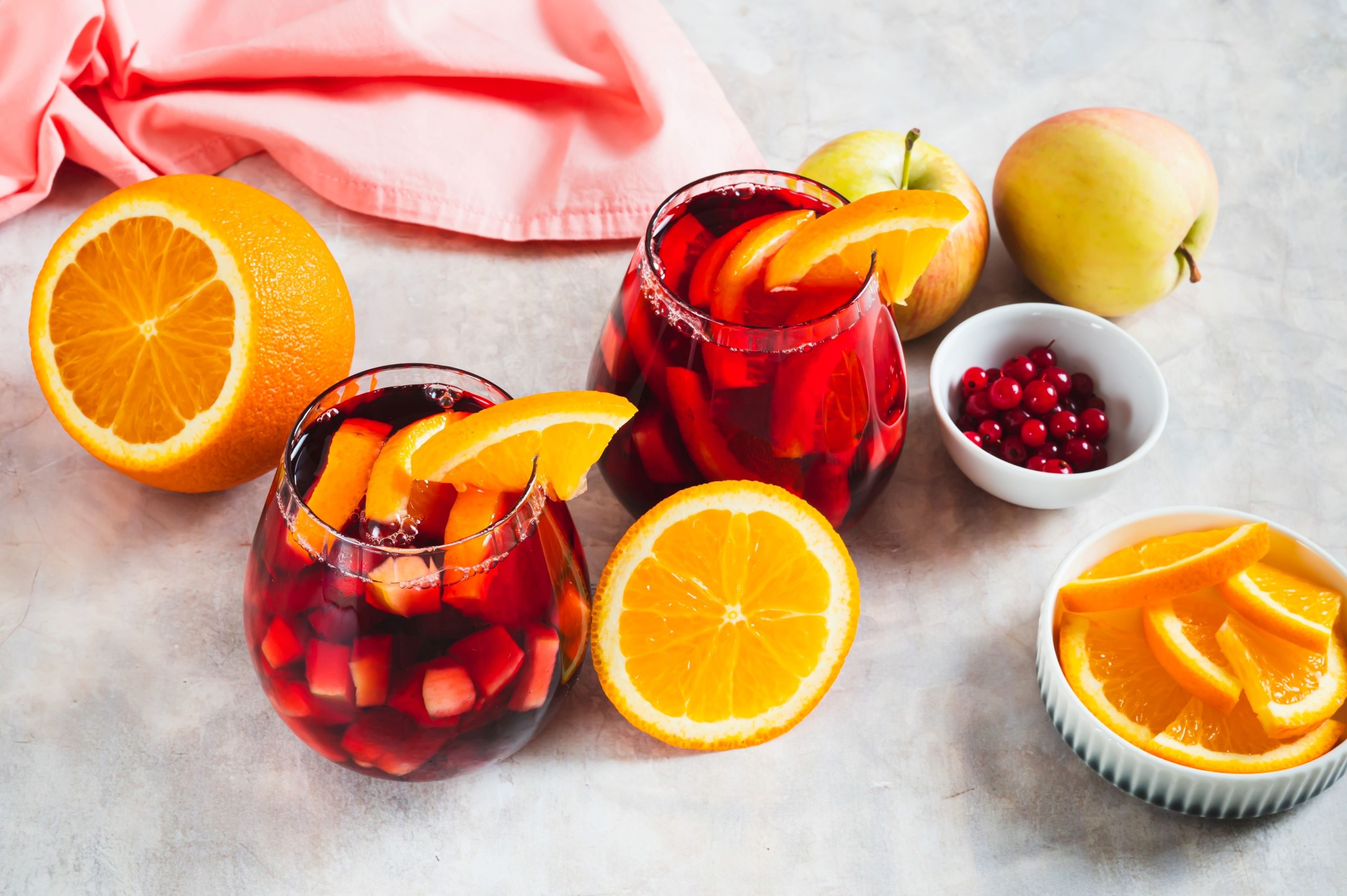 Dvě sklenice červené sangrie s plátky pomeranče, obklopené čerstvými pomeranči, jablky, růžovým ubrusem, miskou brusinek a kousky pomeranče.
