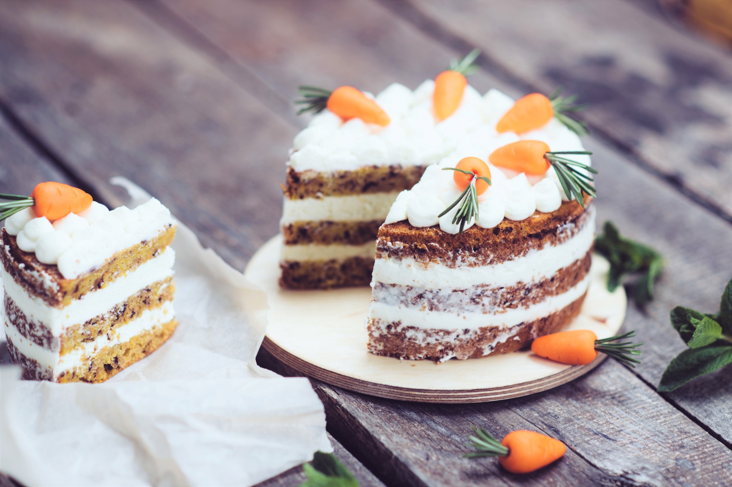 Třívrstvý mrkvový dort s krémovou sýrovou polevou, zdobený malými mrkvovými ozdobami a snítkami bylinek, jeden plátek položen na papíru na dřevěném stole.