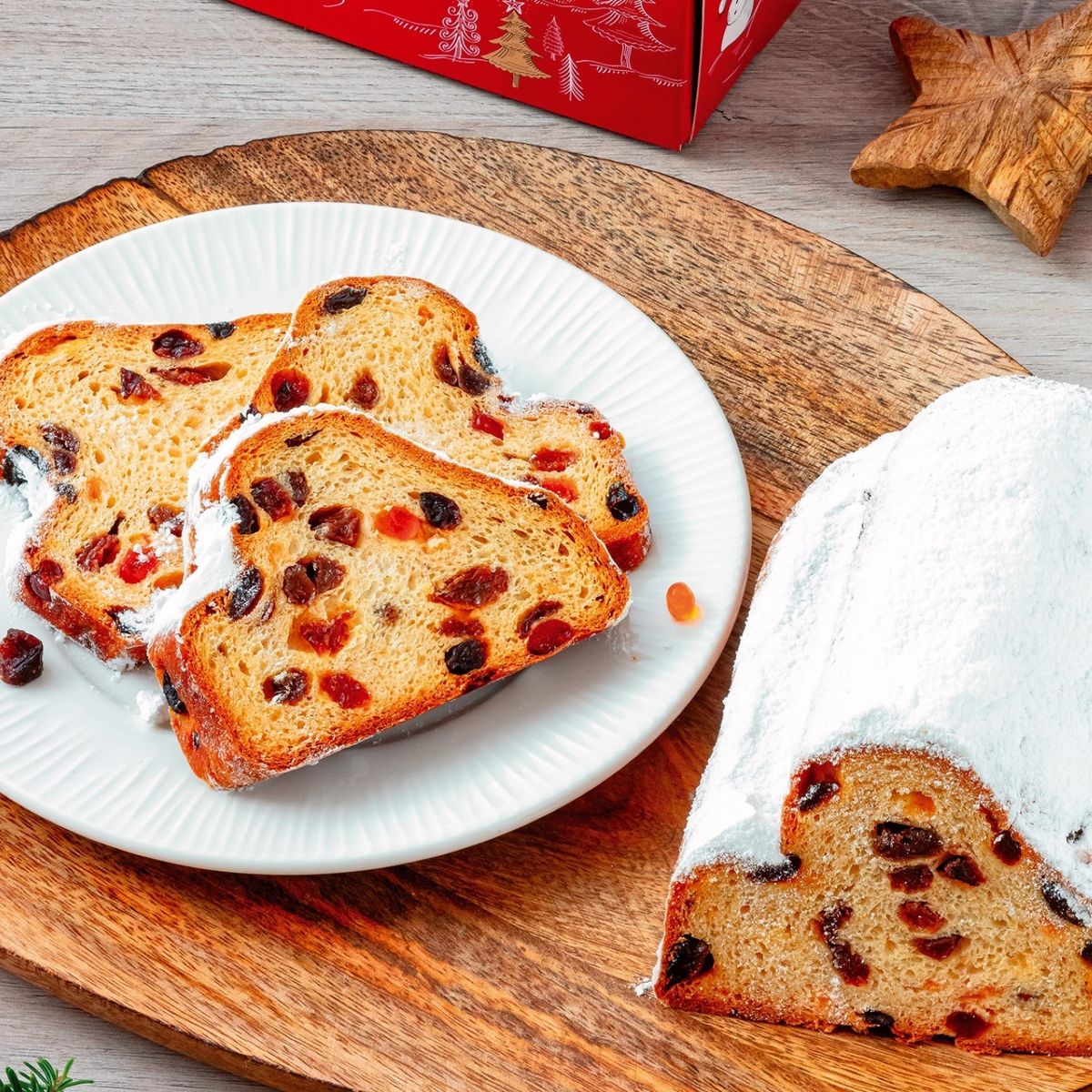 Nakrájená vánoční štola Globus s moučkovým cukrem na bílém talíři a dřevěné desce, obklopená slavnostními dekoracemi.