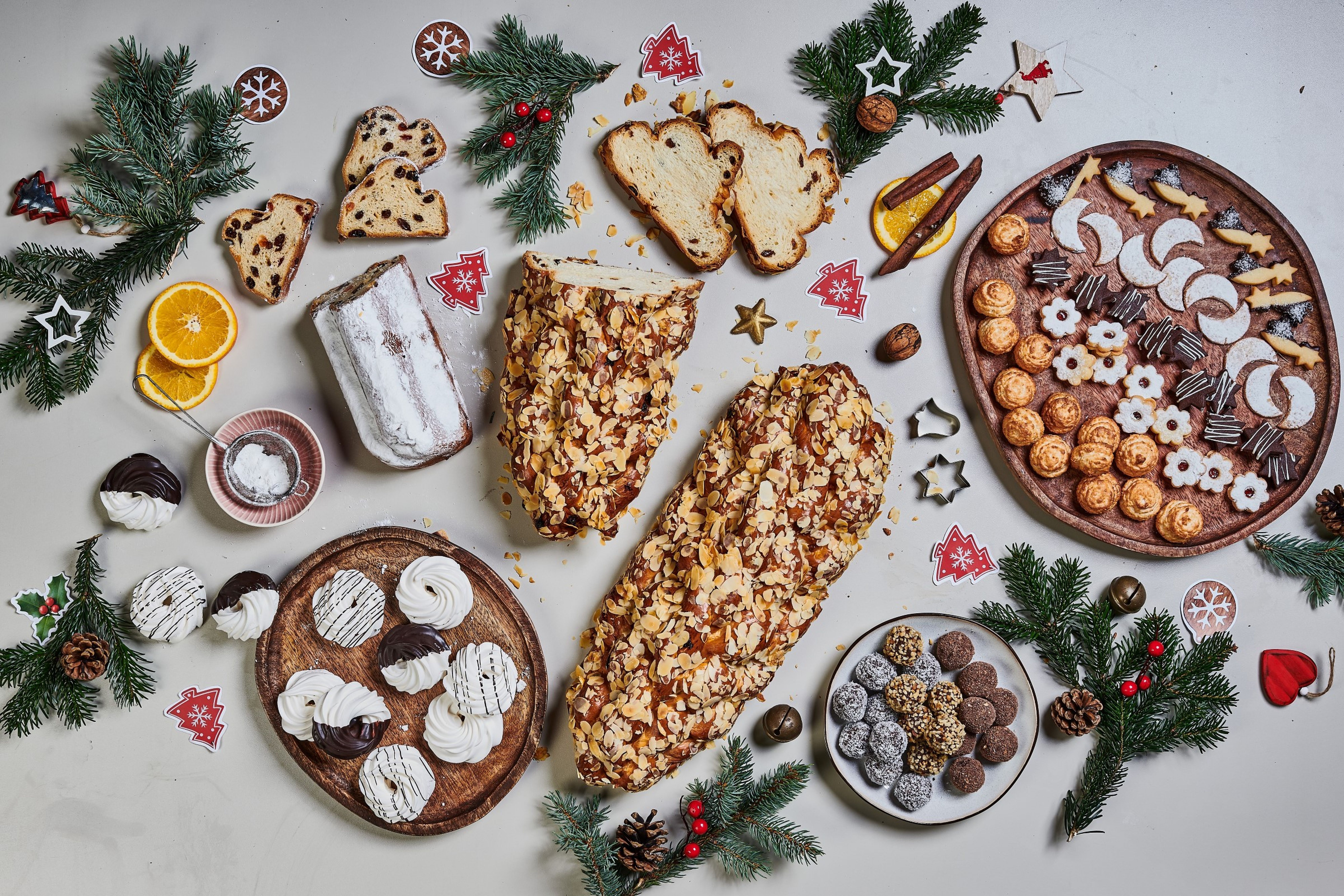 Různé sváteční vánoční pečivo a cukroví, včetně štoly, vánočky, kokosek, lineckého a pusinek, obklopené slavnostními dekoracemi a zelení na bílém povrchu.