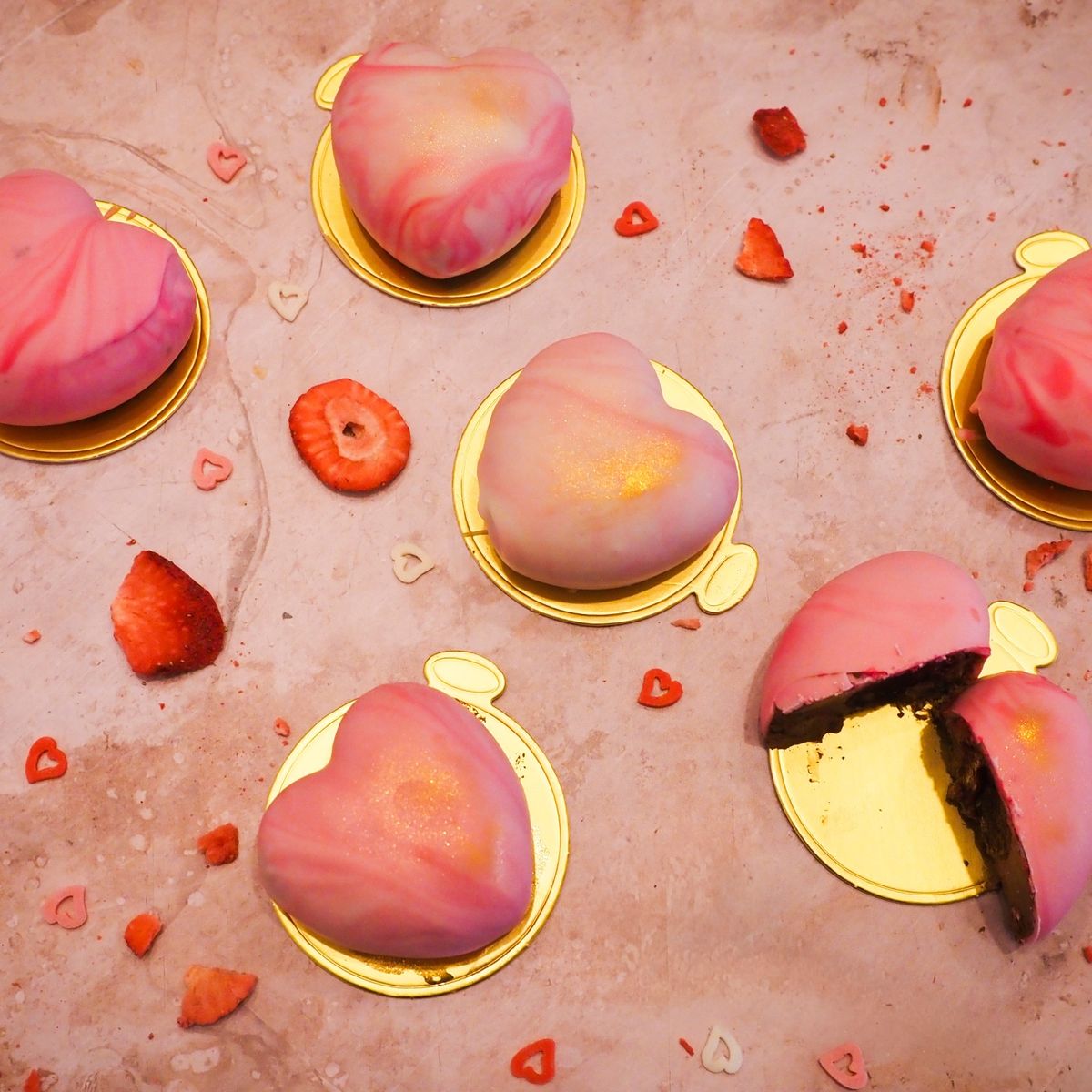 Růžové zákusky ve tvaru srdce na zlatých podložkách, obklopené plátky jahod na mramorovém povrchu. Recept je od Evy z Kitchen Affair.