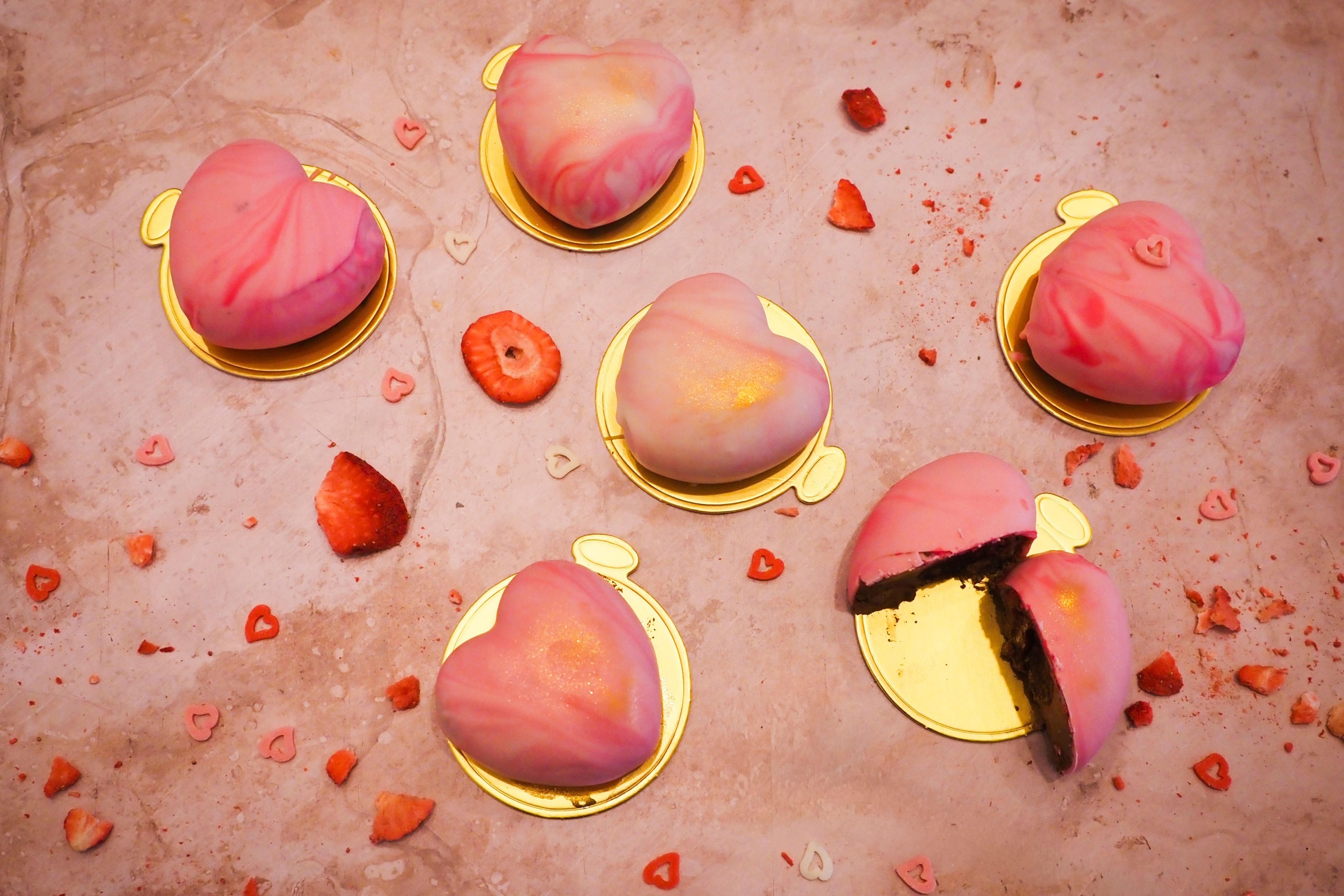 Růžové zákusky ve tvaru srdce na zlatých podložkách, obklopené plátky jahod na mramorovém povrchu. Recept je od Evy z Kitchen Affair.