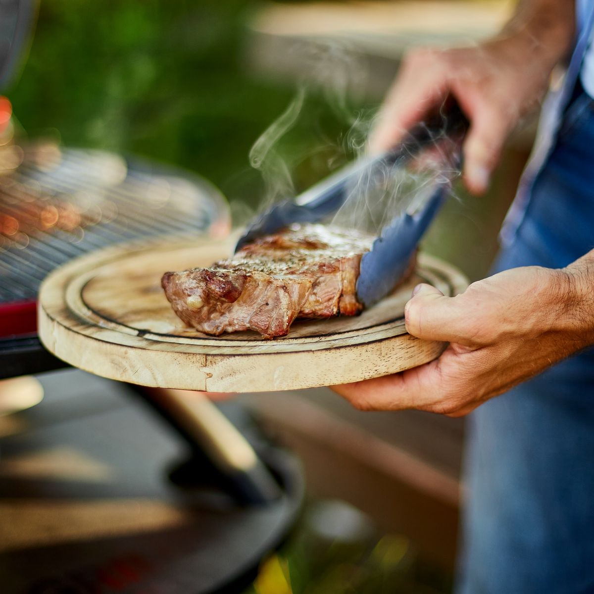 Osoba pokládající grilovaný steak na dřevěný talíř pomocí kleští vedle grilu, v pozadí je vidět kukuřice.