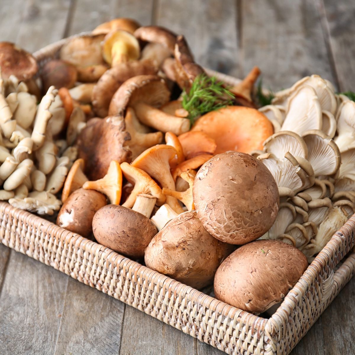 Proutěný podnos plný různých hub, včetně hlívy ústřičné, shiitake a žampionů, na rustikálním dřevěném stole.