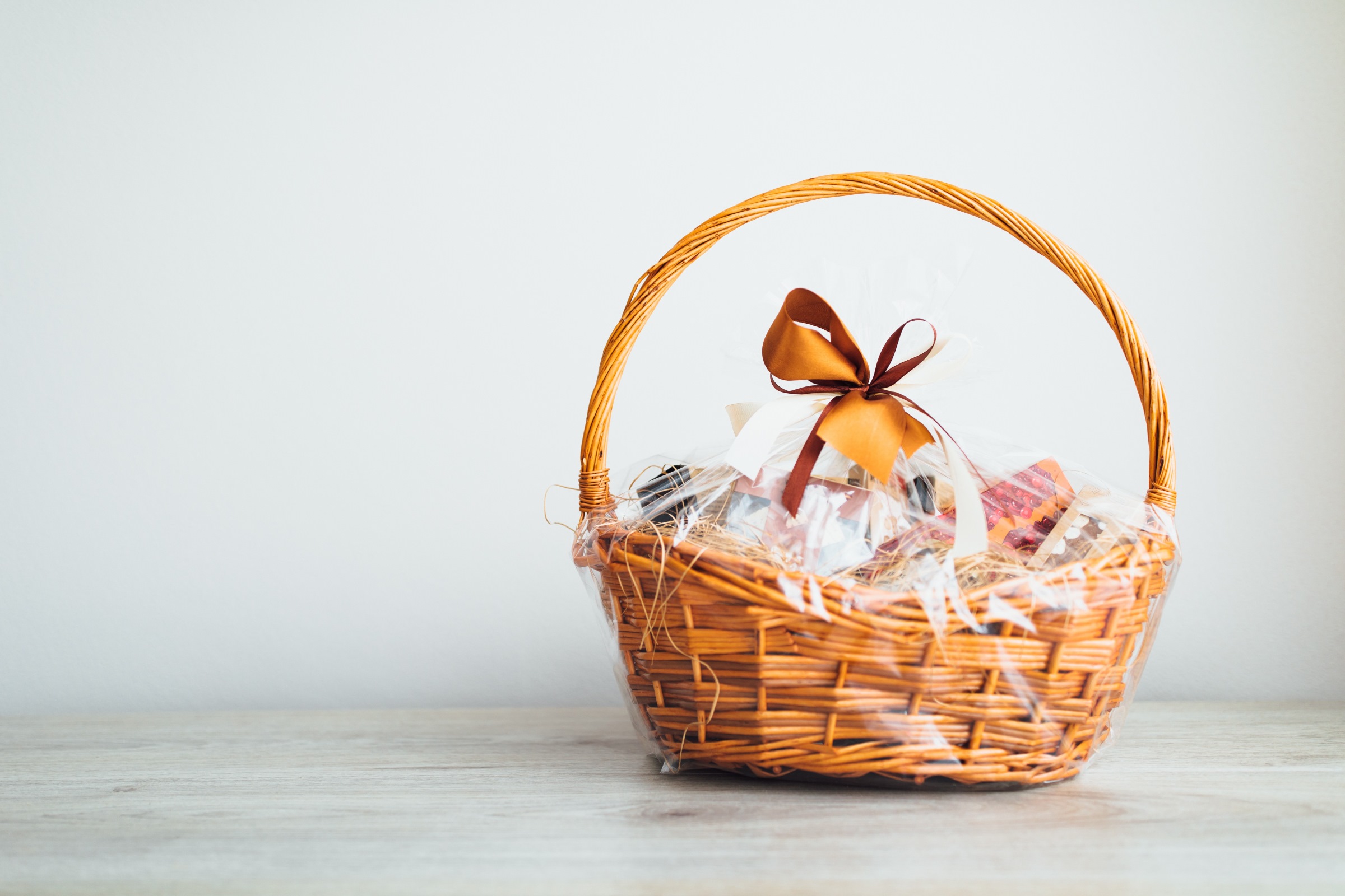 Proutěný dárkový koš zabalený v celofánu, převázaný hnědou stuhou, umístěný na dřevěné ploše před jednobarevným bílým pozadím.
