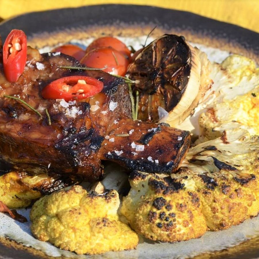 Grilovaná hovězí žebírka zdobená červeným chilli a bylinkami, podávaná s pečeným květákem a opečenou limetkou na rustikálním talíři.