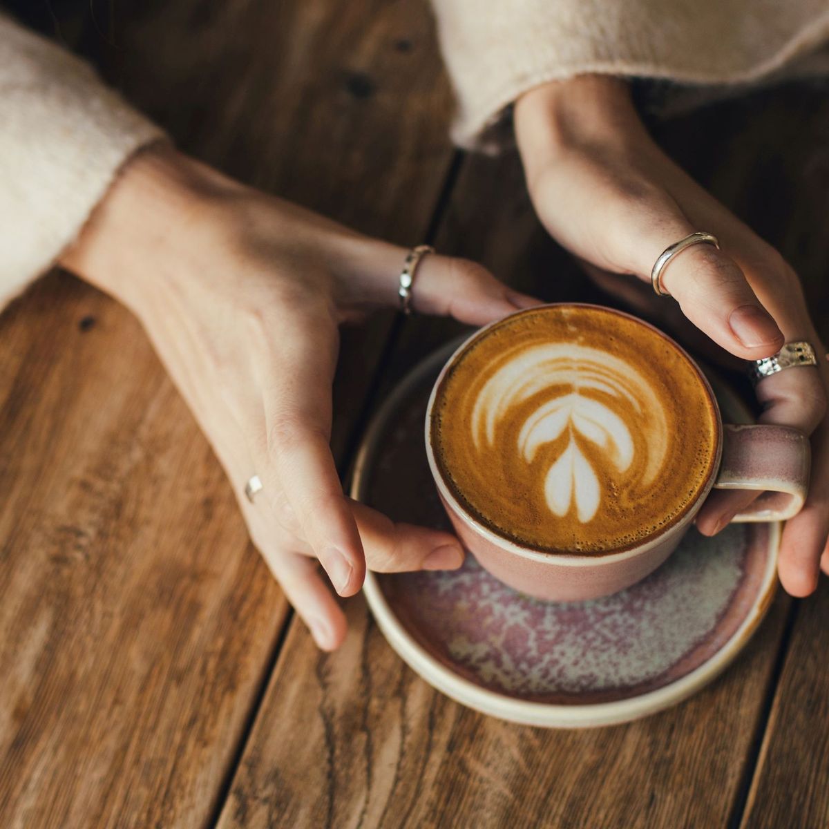 Ruce držící šálek latté s pěnovým vzorem ve tvaru listu na dřevěném stole.
