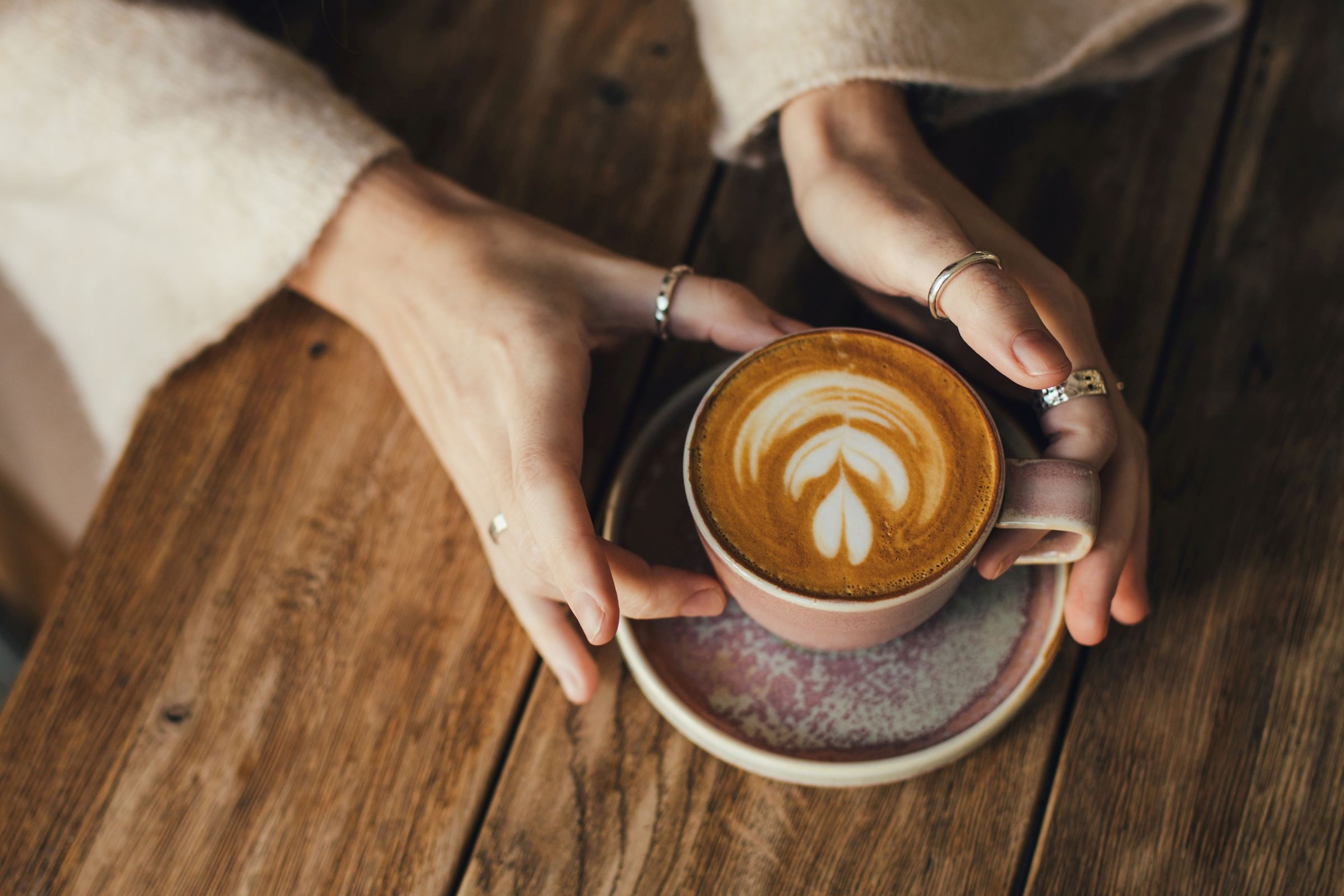 Ruce držící šálek latté s pěnovým vzorem ve tvaru listu na dřevěném stole.