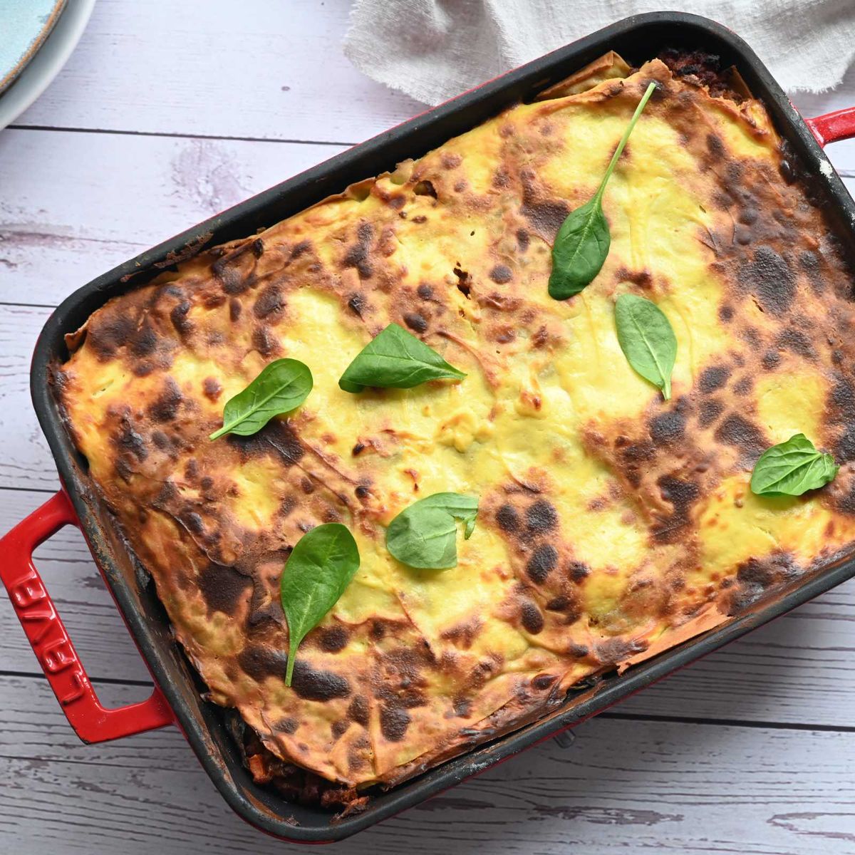 Zapečené lasagne se zlatavou sýrovou krustou ozdobené čerstvými listy bazalky v červené obdélníkové zapékací míse na bílém dřevěném stole. Recept připravil Vojta Poštulka