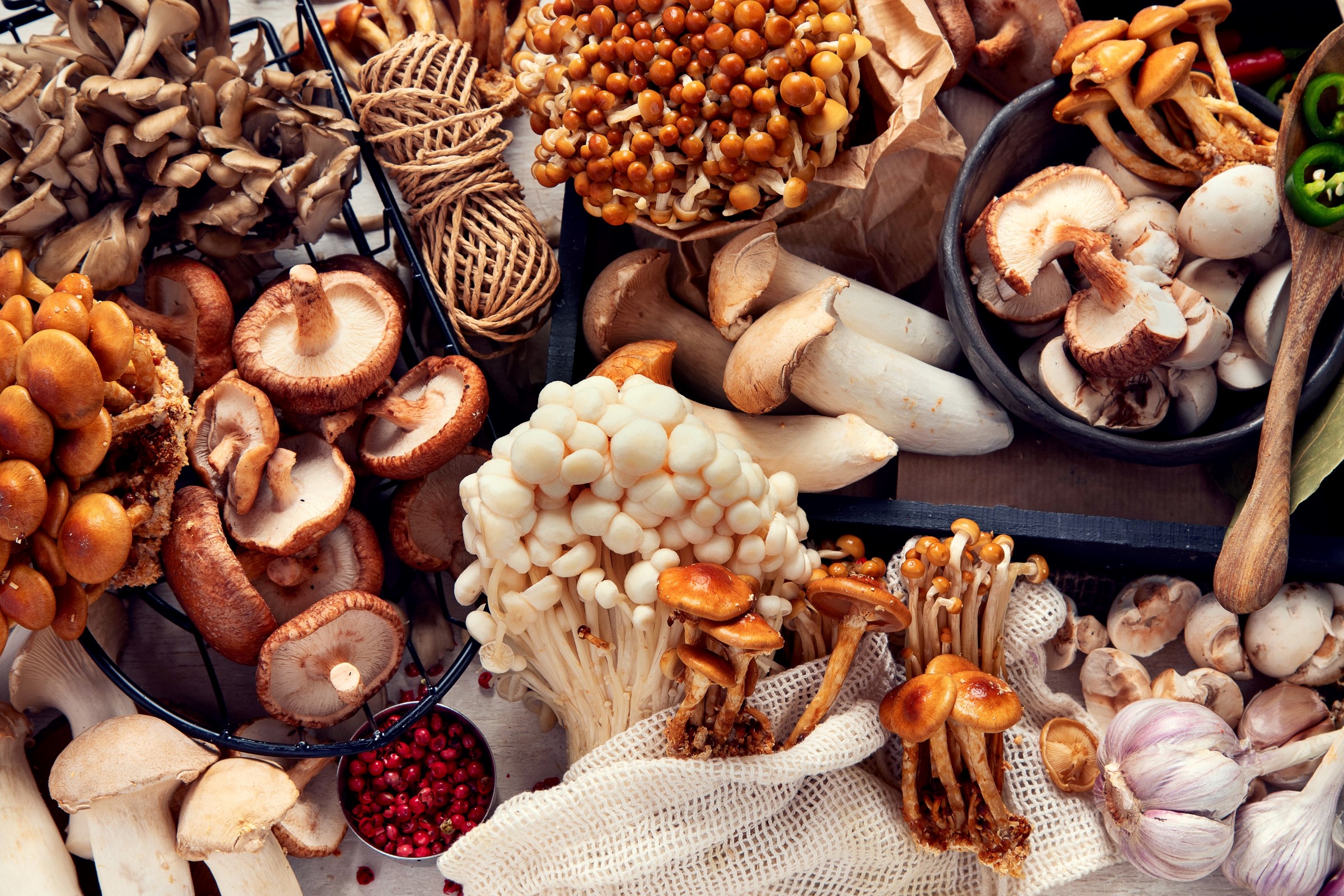 Různé druhy hub, včetně shiitake, enoki a dalších, jsou na dřevěné desce naskládány spolu s česnekem a červeným pepřem.