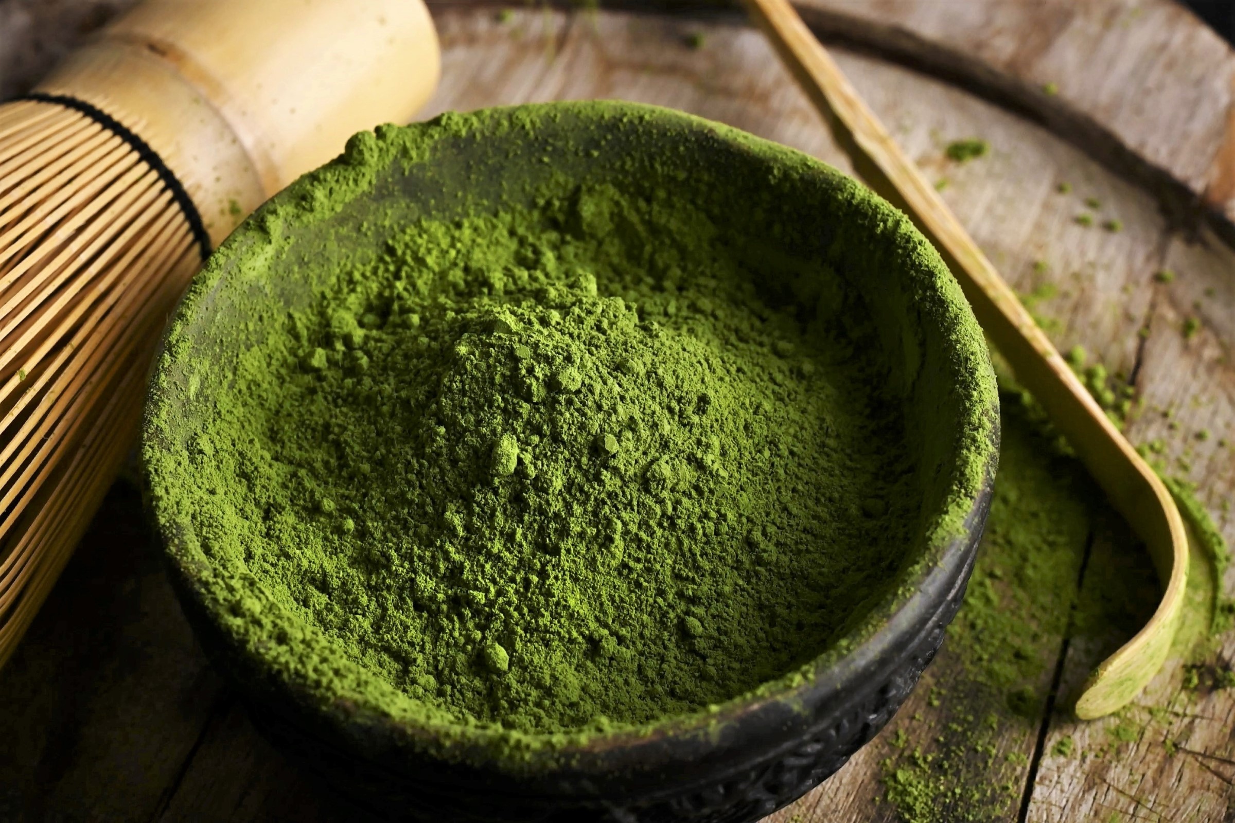 Miska s jasně zeleným práškem matcha, k níž patří bambusová metlička a lžička, položená na dřevěné podložce.