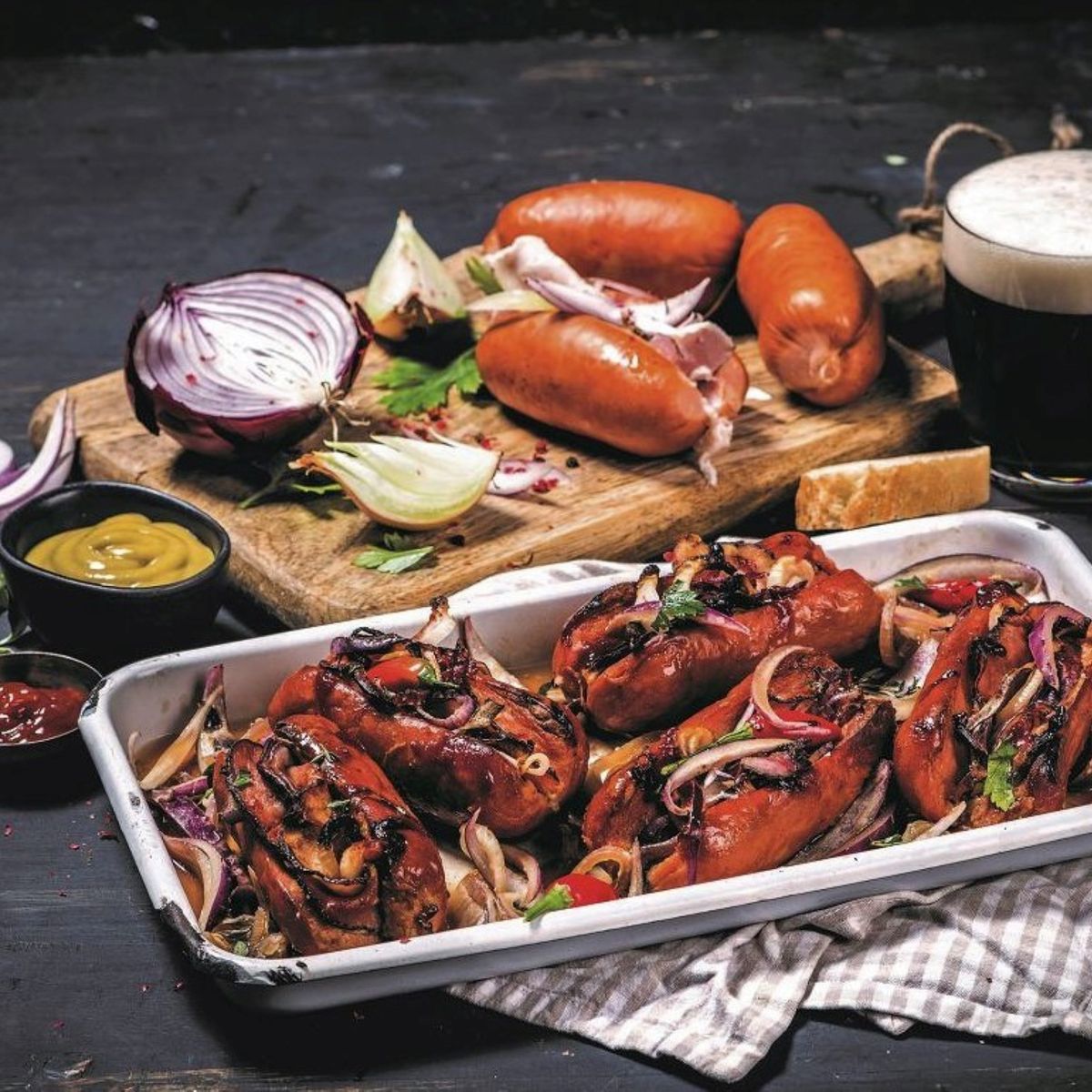 Opečené špekáčky na talíři s cibulí a bylinkami, vedle dřevěné desky se syrovými špekáčky, cibulí, hořčicí a sklenicí černého piva.