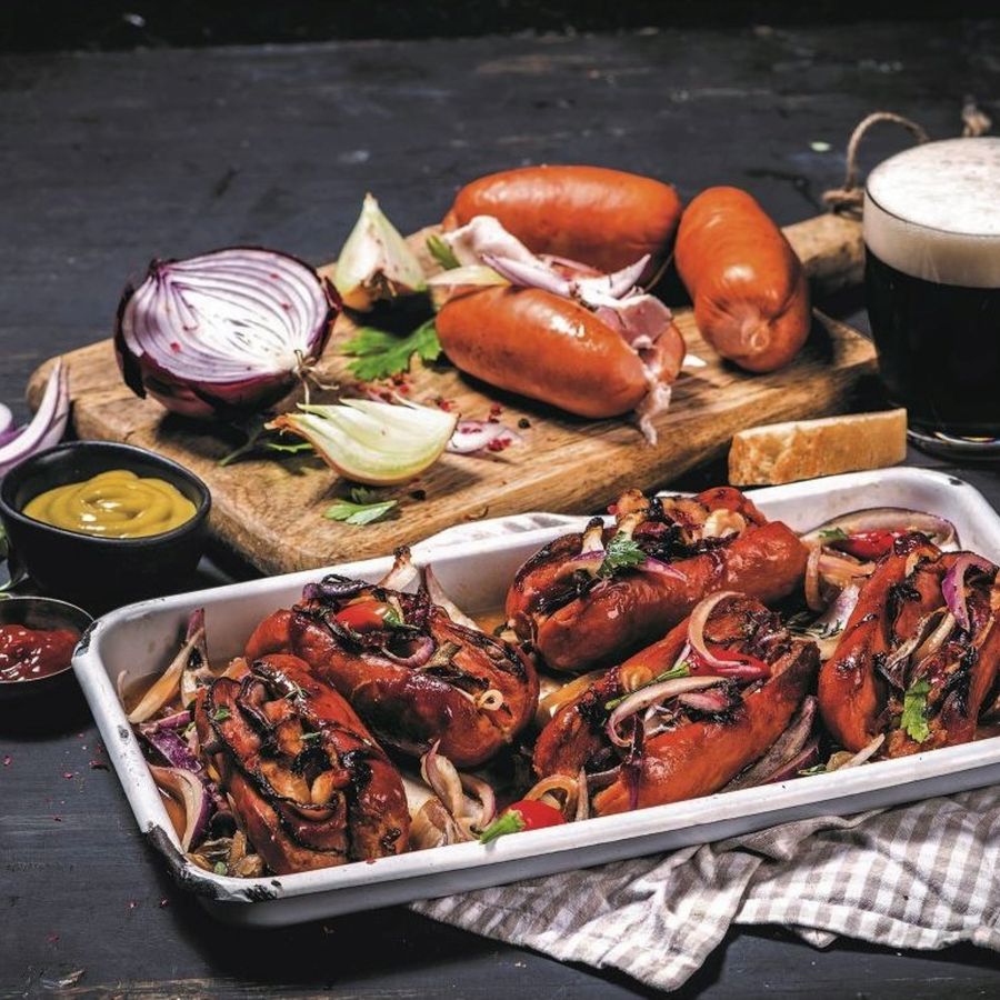 Opečené špekáčky na talíři s cibulí a bylinkami, vedle dřevěné desky se syrovými špekáčky, cibulí, hořčicí a sklenicí černého piva.