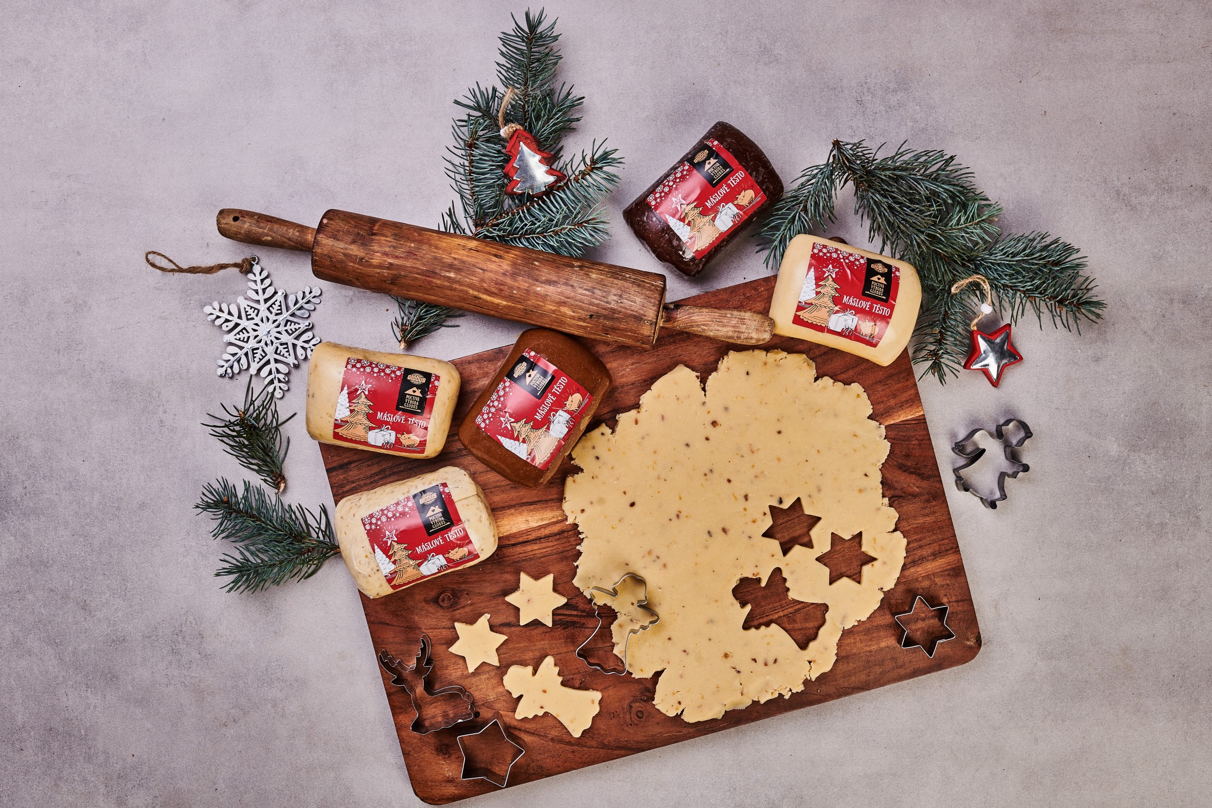 Dřevěná deska s rozváleným těstem na cukroví, vykrajovátky ve tvaru hvězdiček a stromečků, váleček, bloky různých těst na vánoční cukroví, borovicové větvičky a sněhové vločky jako ozdoba.
