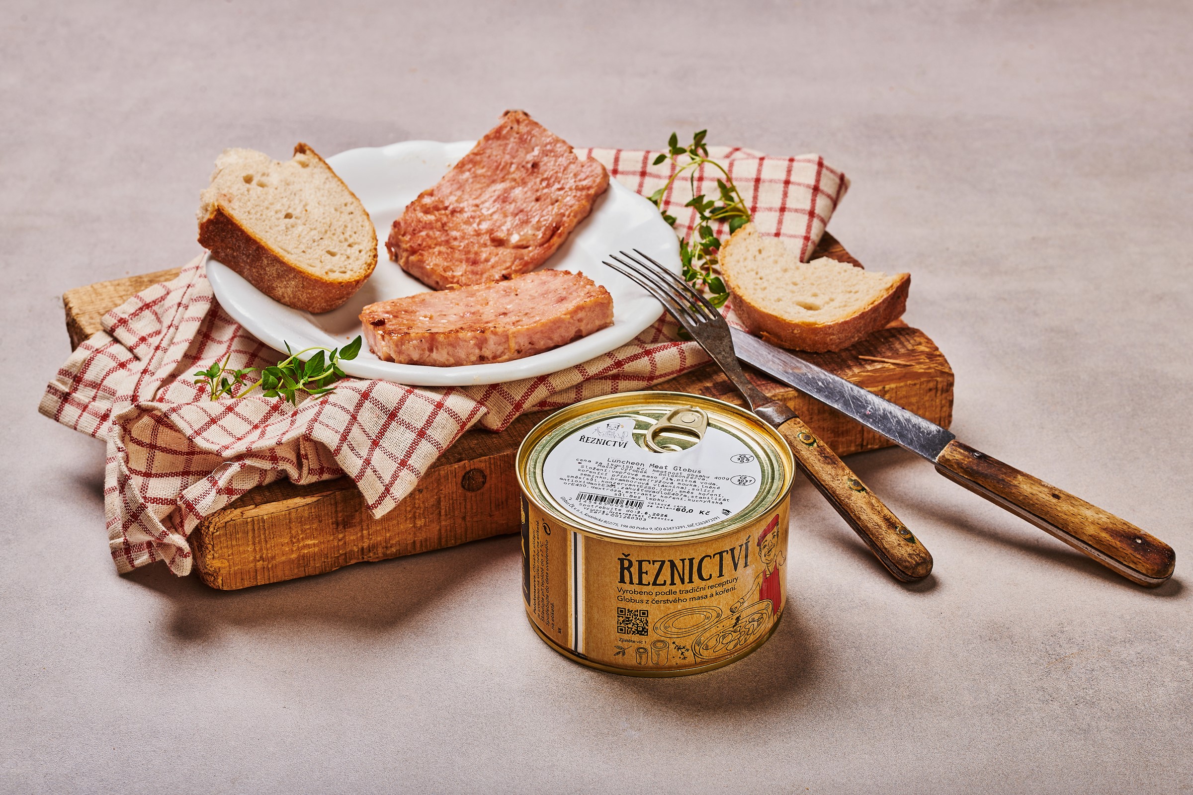 Nakrájený Luncheon Meat Globus a chléb na talíři s bylinkami, vedle polokonzervy Globus, s nožem a vidličkou na dřevěné desce pokryté kostkovanou utěrkou.