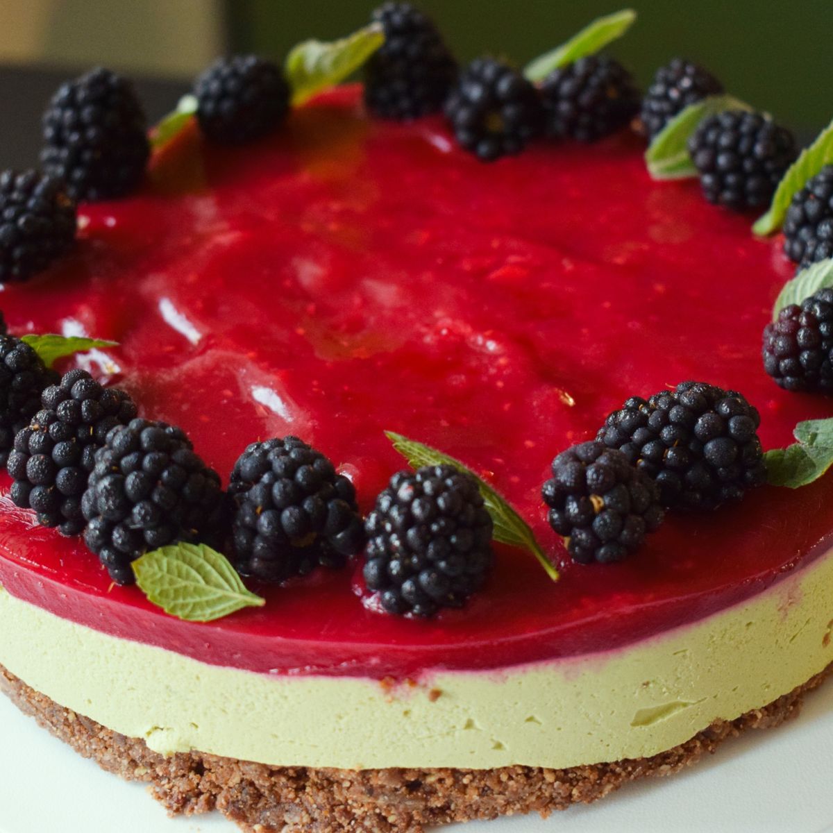 Cheesecake s matcha korpusem a malinovou vrstvou, ozdobený ostružinami a lístky máty, na bílém talíři. Recept připravily Ségry v kuchyni.