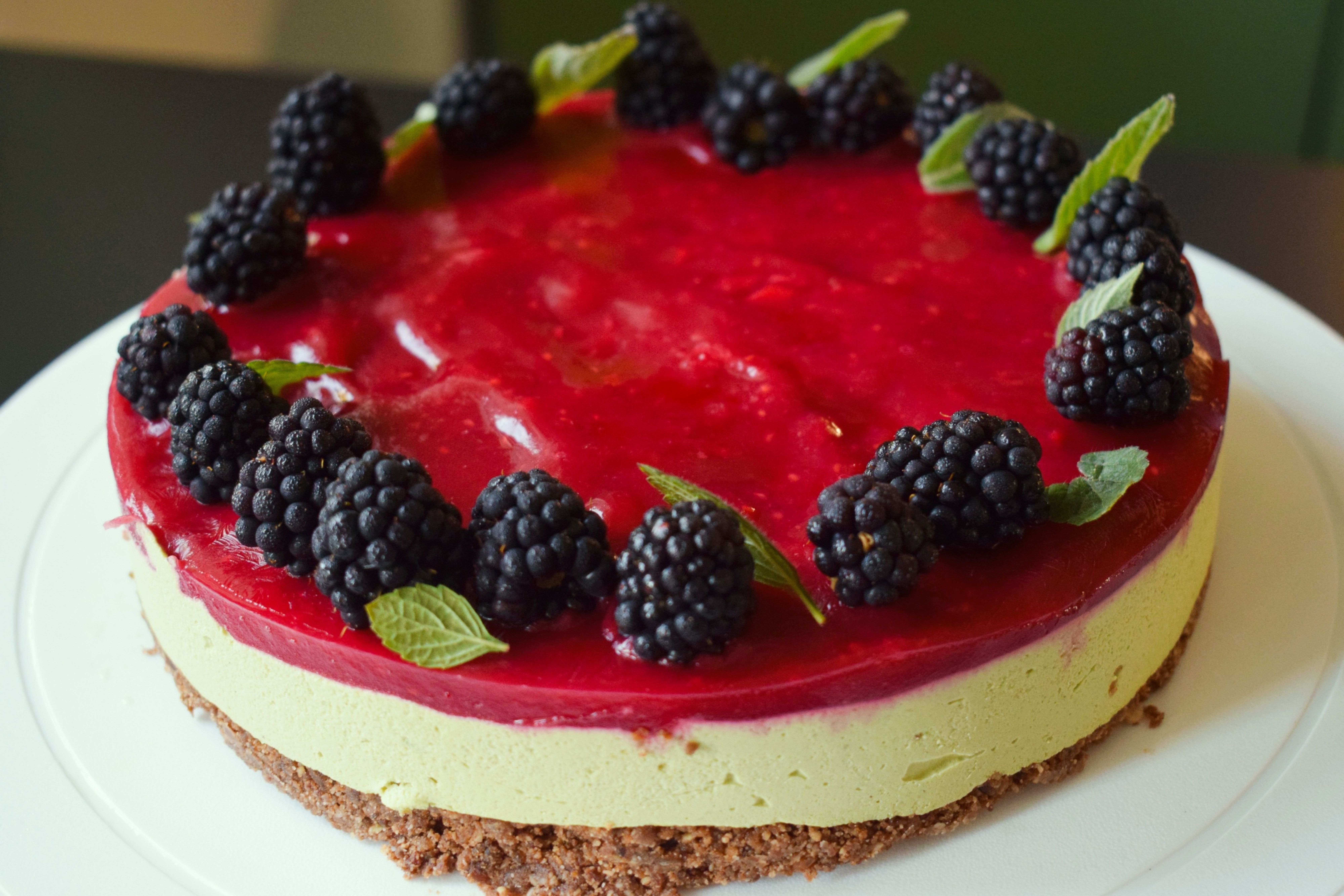 Cheesecake s matcha korpusem a malinovou vrstvou, ozdobený ostružinami a lístky máty, na bílém talíři. Recept připravily Ségry v kuchyni.