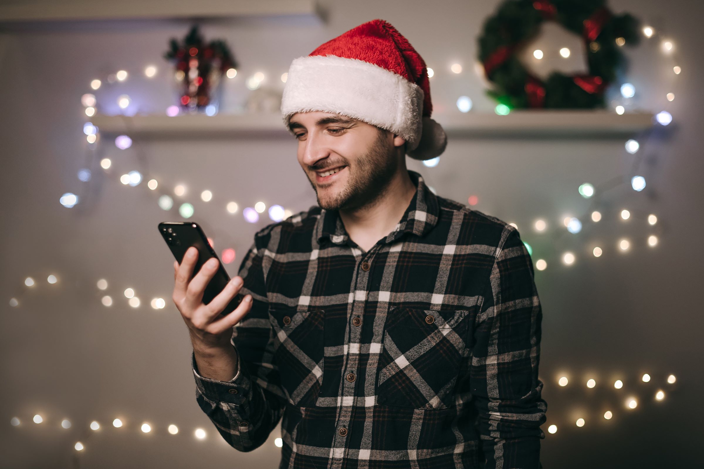 Muž v čepici Santa Clause se usmívá na svůj telefon, v pozadí jsou sváteční světla a věnec.