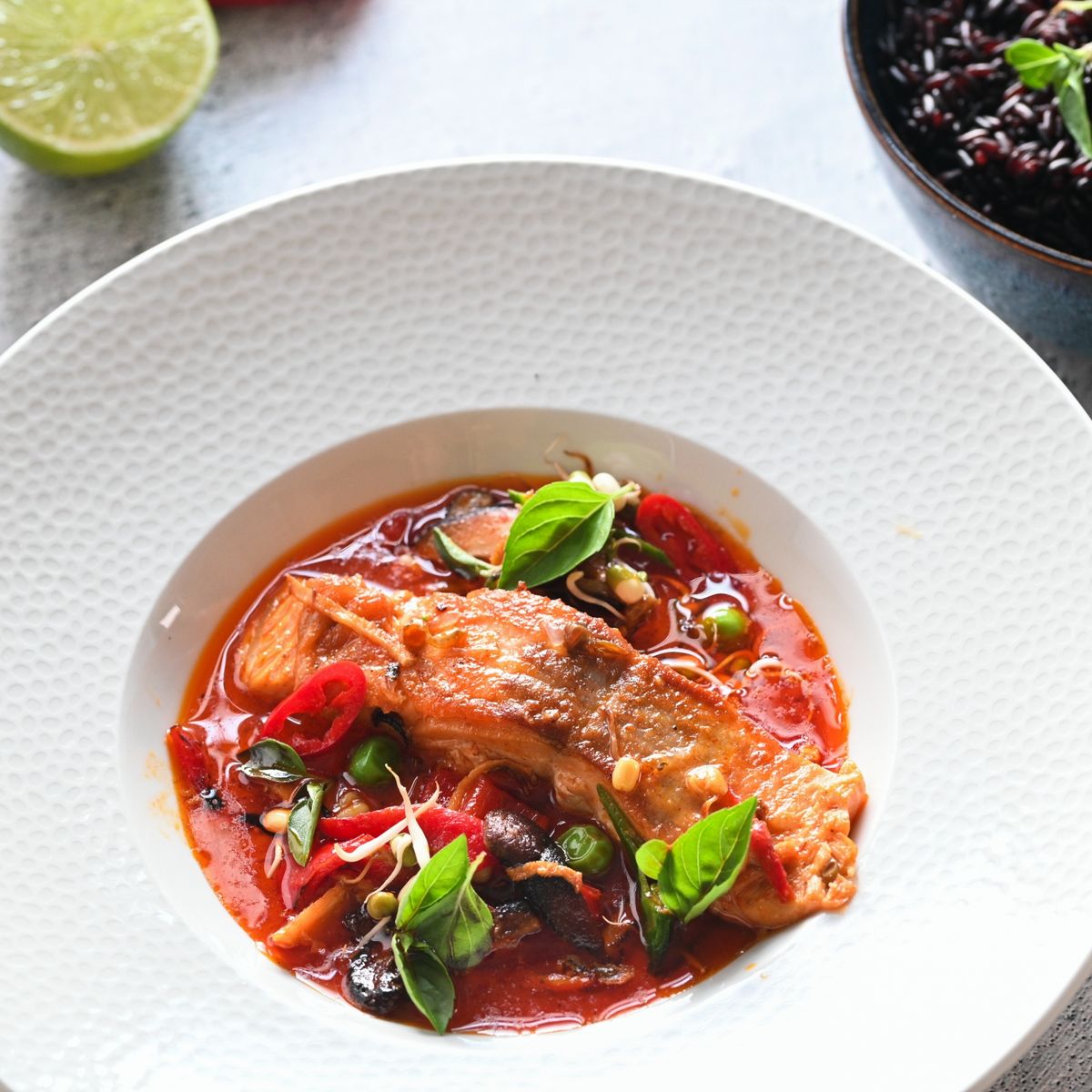 Talíř s červeným curry s lososem, ozdobená čerstvými listy bazalky a nakrájenými chilli papričkami, vedle miska s černou rýží a půlka limetky. Recept připravil Vojta Poštulka.