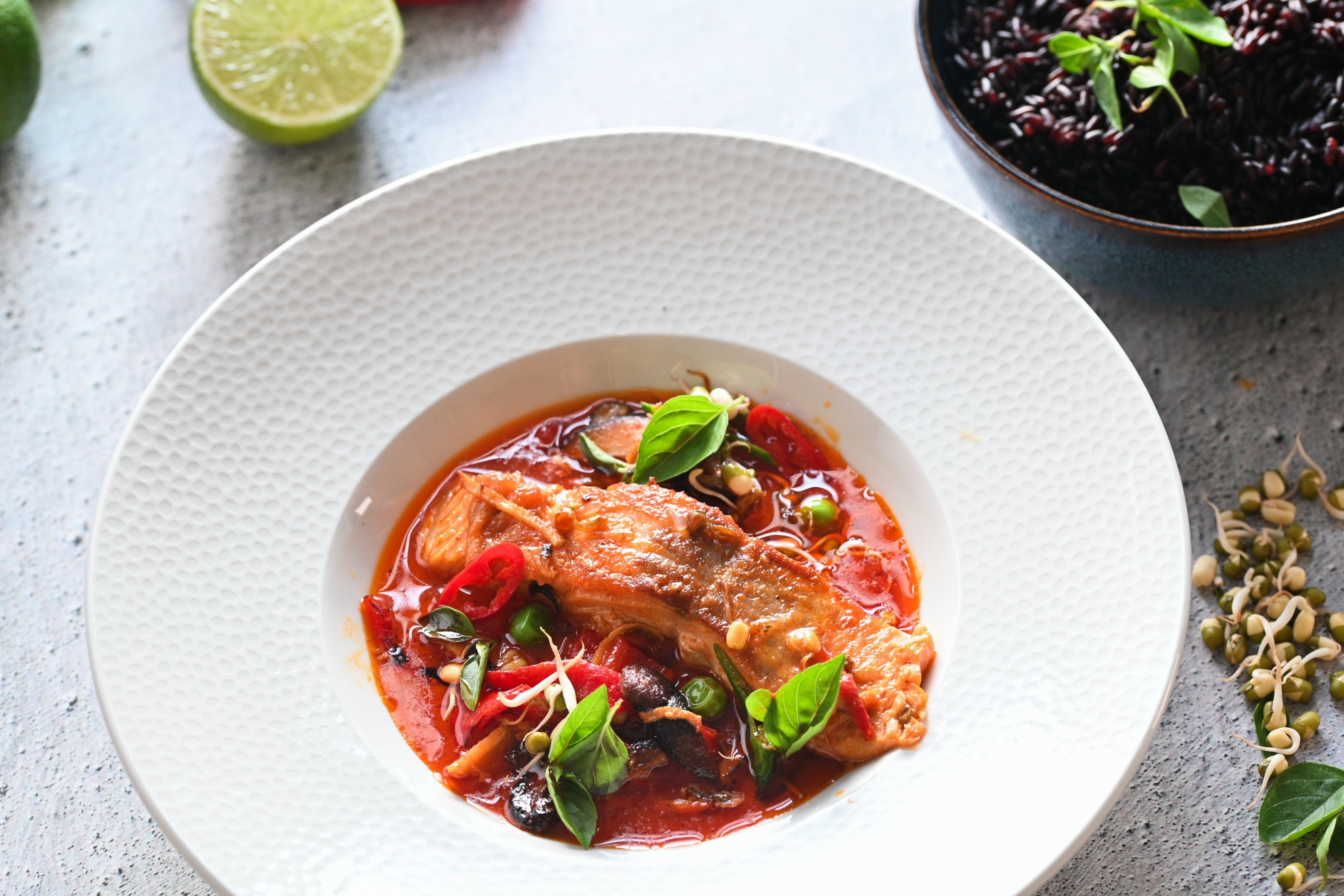 Talíř s červeným curry s lososem, ozdobená čerstvými listy bazalky a nakrájenými chilli papričkami, vedle miska s černou rýží a půlka limetky. Recept připravil Vojta Poštulka.